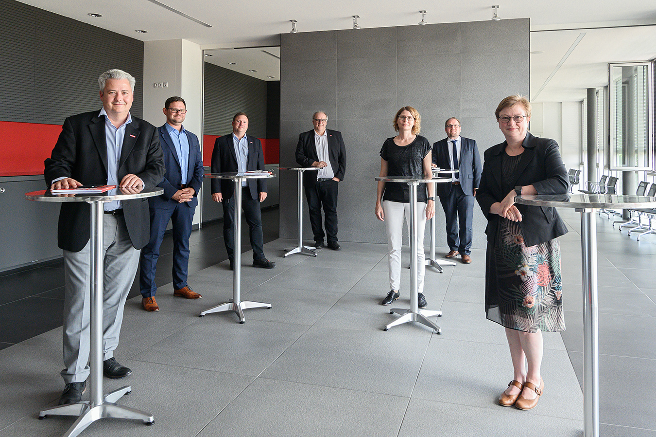 Eine Gruppe von sieben Personen in Business-Kleidung steht in einem modernen Raum mit hohen Tischen an der Hochschule Coburg. Der Raum ist in Grau und Rot gehalten und wird auf einer Seite durch große Fenster erhellt. Die Gruppe lächelt und nimmt an einem scheinbar zwanglosen Treffen teil.