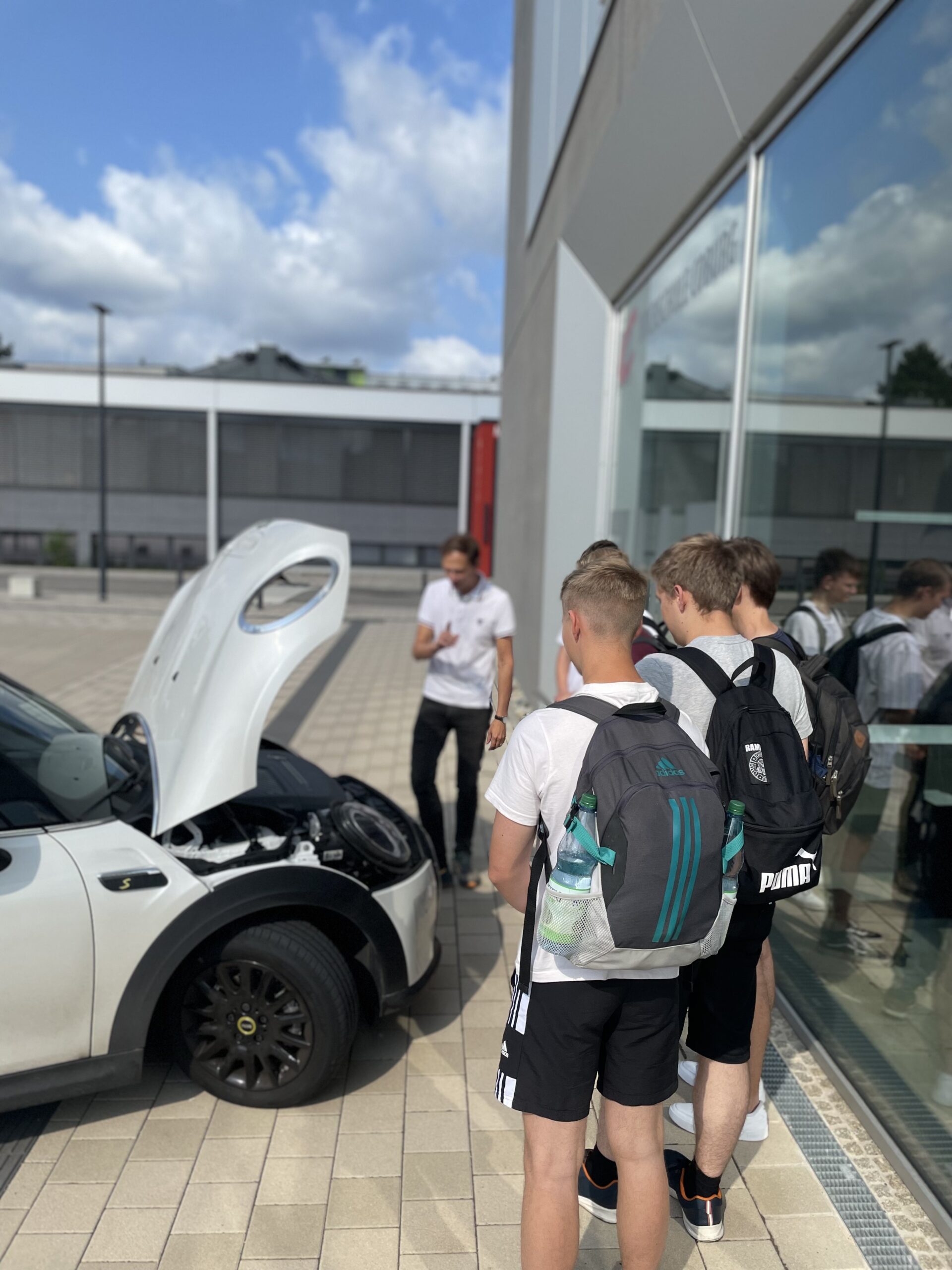 Eine Gruppe junger Leute mit Rucksäcken versammelt sich um ein weißes Auto mit geöffneter Motorhaube. Daneben erzählt eine Person im weißen Hemd eifrig etwas über die Hochschule Coburg. Die Szene spielt sich im Freien an einem sonnigen Tag ab.