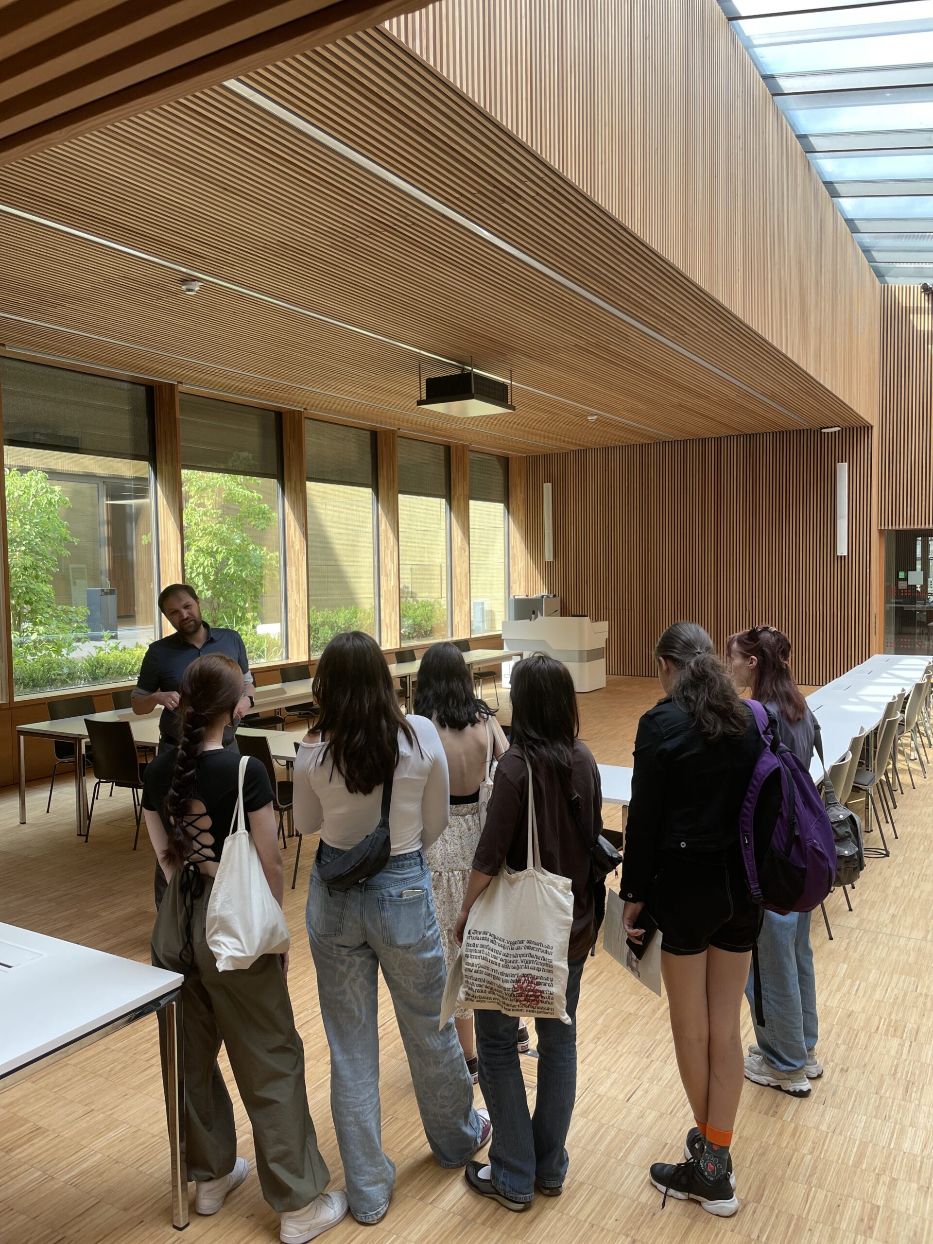 Eine Gruppe von Menschen steht in einem geräumigen, holzgetäfelten Raum mit großen Fenstern und Oberlicht in der Hochschule Coburg. Sie hören gespannt einem Redner am Tisch zu, während im Hintergrund ordentlich aufgestellte Tische und Stühle stehen.