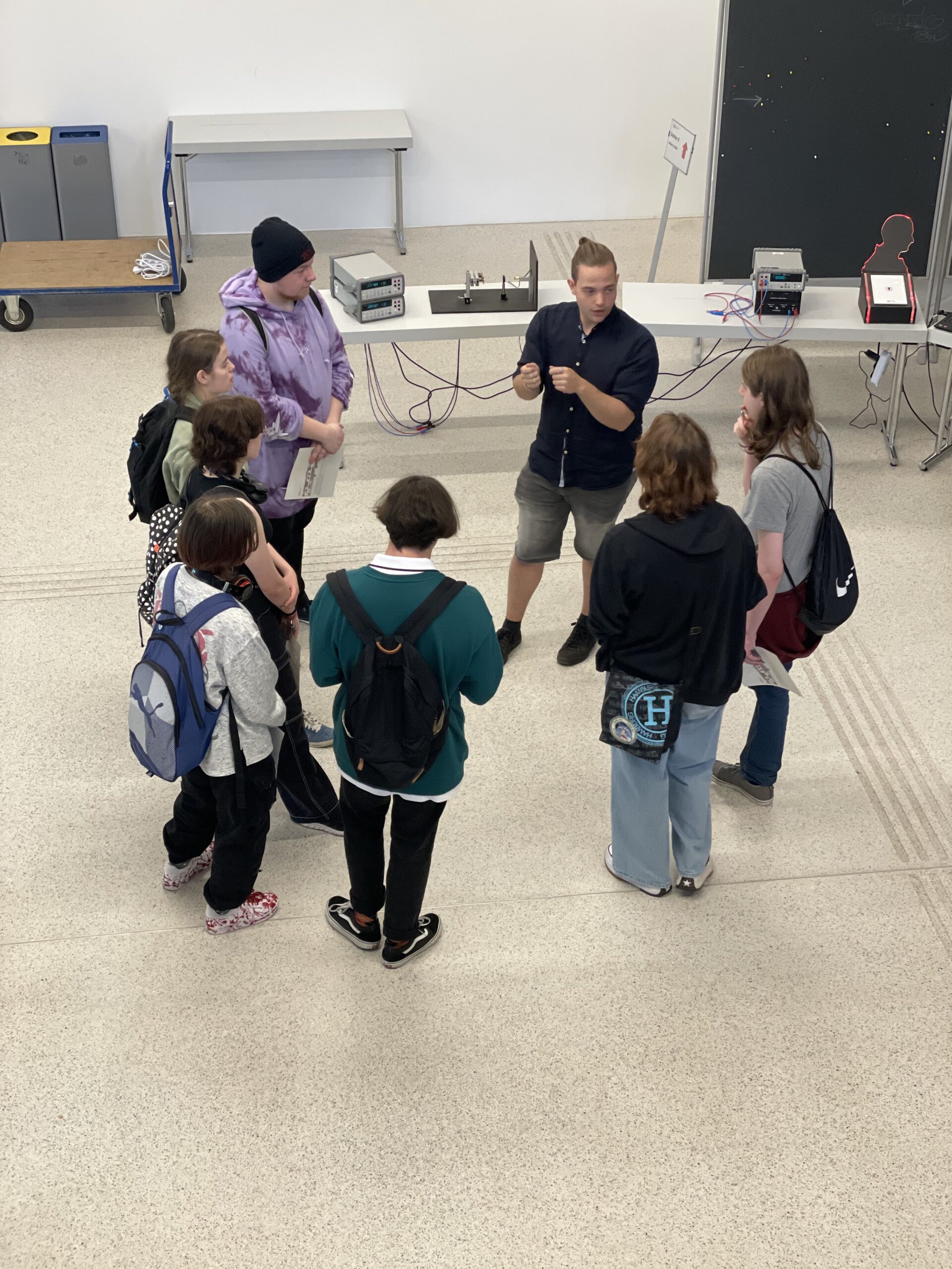 Eine Gruppe von Menschen versammelt sich in einem Innenbereich der Hochschule Coburg in einem Kreis und hört aufmerksam zu, wie jemand ein Konzept erklärt. Im Hintergrund sind elektronische Geräte auf einem Tisch angeordnet, was vor dem hellen Boden die Unterrichtsatmosphäre verstärkt.