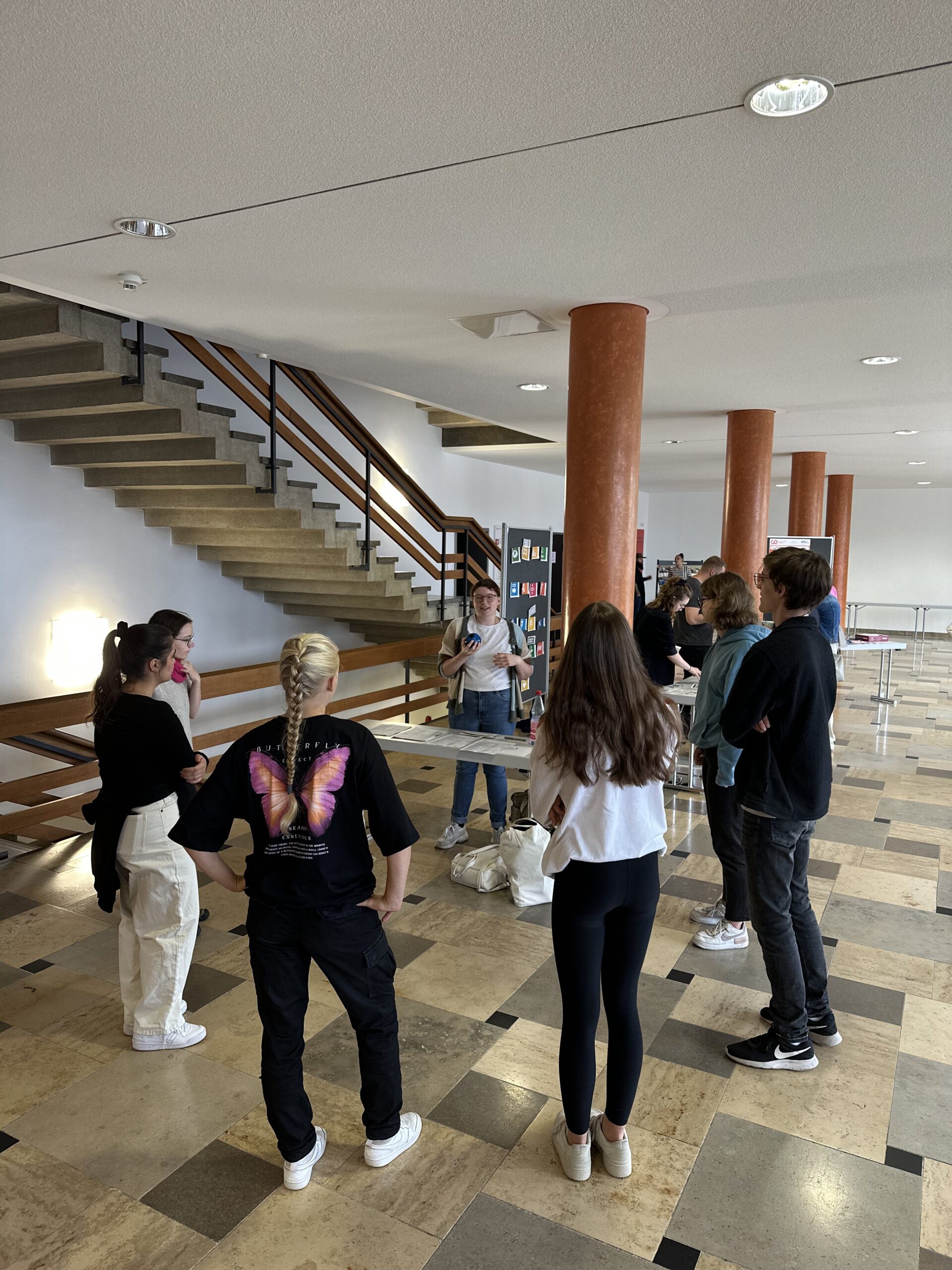 Eine Gruppe von Menschen steht im Innenbereich der Hochschule Coburg im Kreis und hört einem Vortragenden zu. Sie versammeln sich in einem geräumigen Bereich mit einer Treppe im Hintergrund. Die meisten sind leger gekleidet, und eine Person trägt ein schwarzes Hemd mit einem großen Schmetterlingsmuster auf dem Rücken.