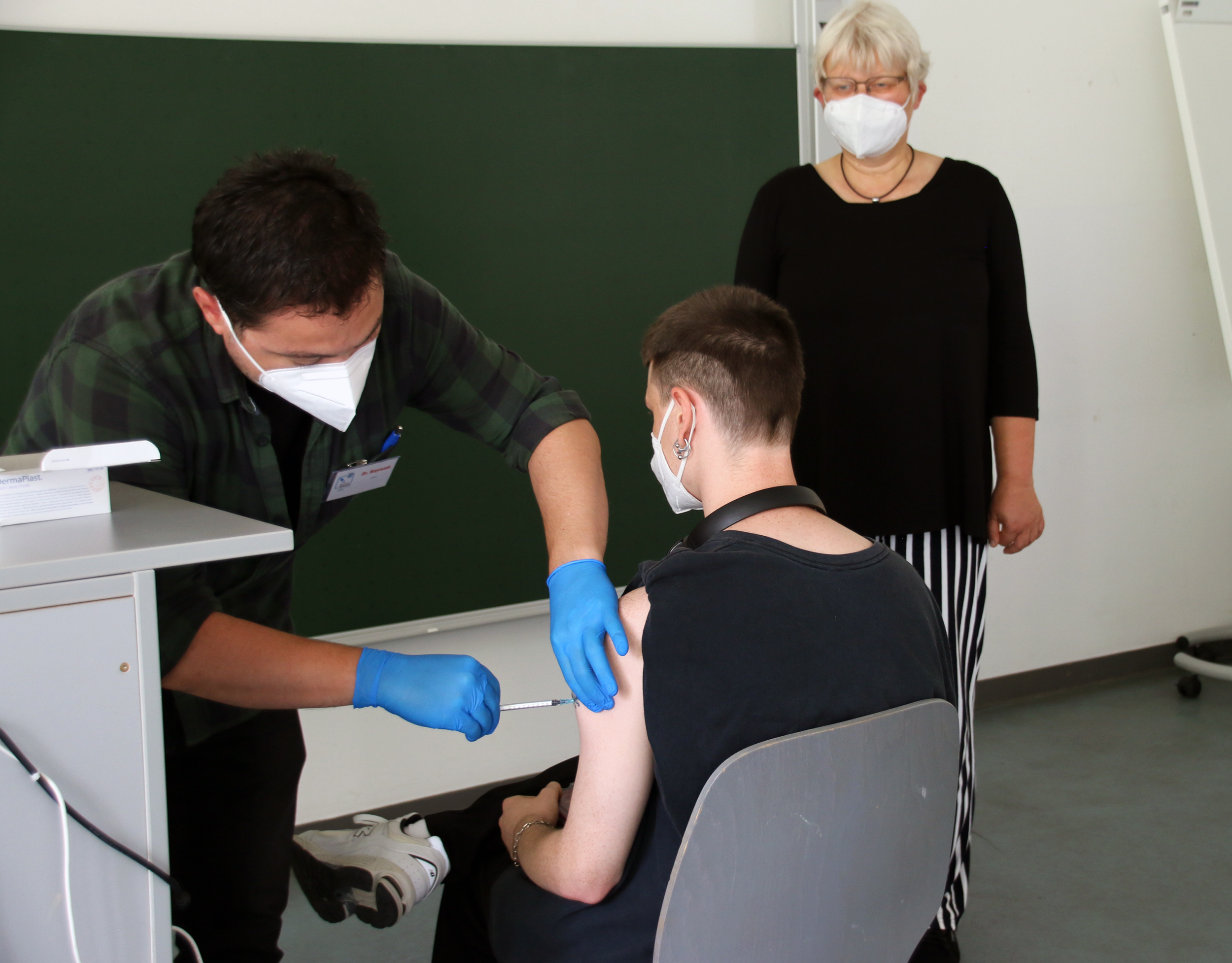 Eine Person mit Maske und Handschuhen verabreicht einer sitzenden Person an der Hochschule Coburg eine Impfstoffinjektion. Eine weitere Person steht in der Nähe und beobachtet den Vorgang. Alle sind maskiert und im Hintergrund ist eine grüne Tafel zu sehen.