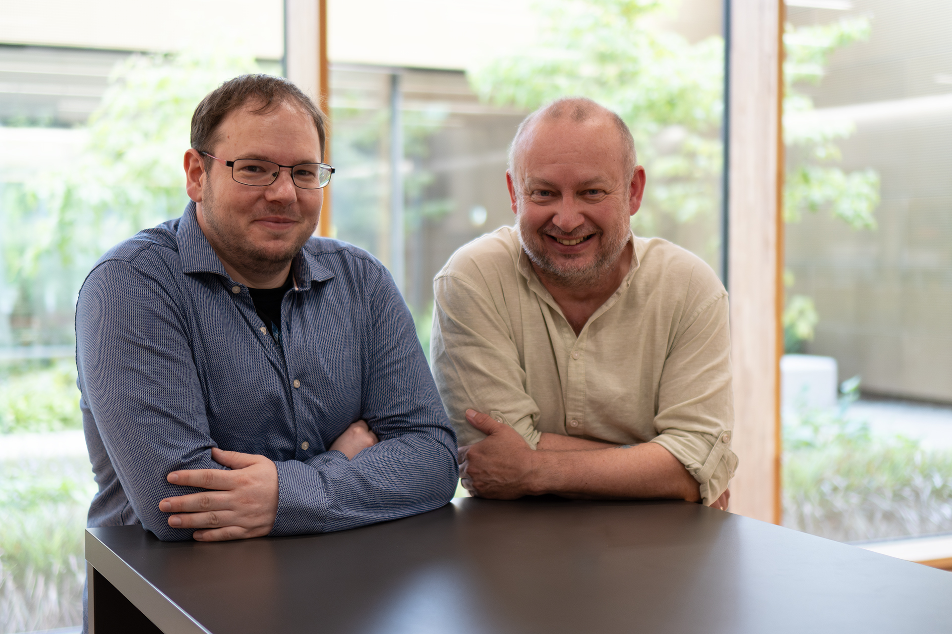 Two men are smiling and leaning on a black table in front of a large window, showcasing the lush greenery outside. Indoors at Hochschule Coburg, one wears a blue shirt and glasses, the other a light beige shirt, both radiating camaraderie.