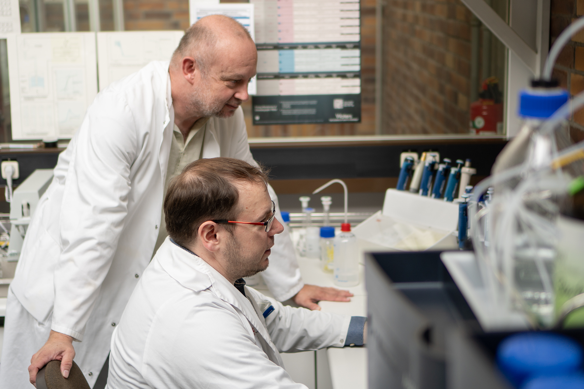 Two scientists in white lab coats collaborate in a Hochschule Coburg laboratory. One stands, examining the computer screen, while the other sits, deeply focused on the monitor. Lab equipment and charts surround them, creating an atmosphere of innovation and discovery.