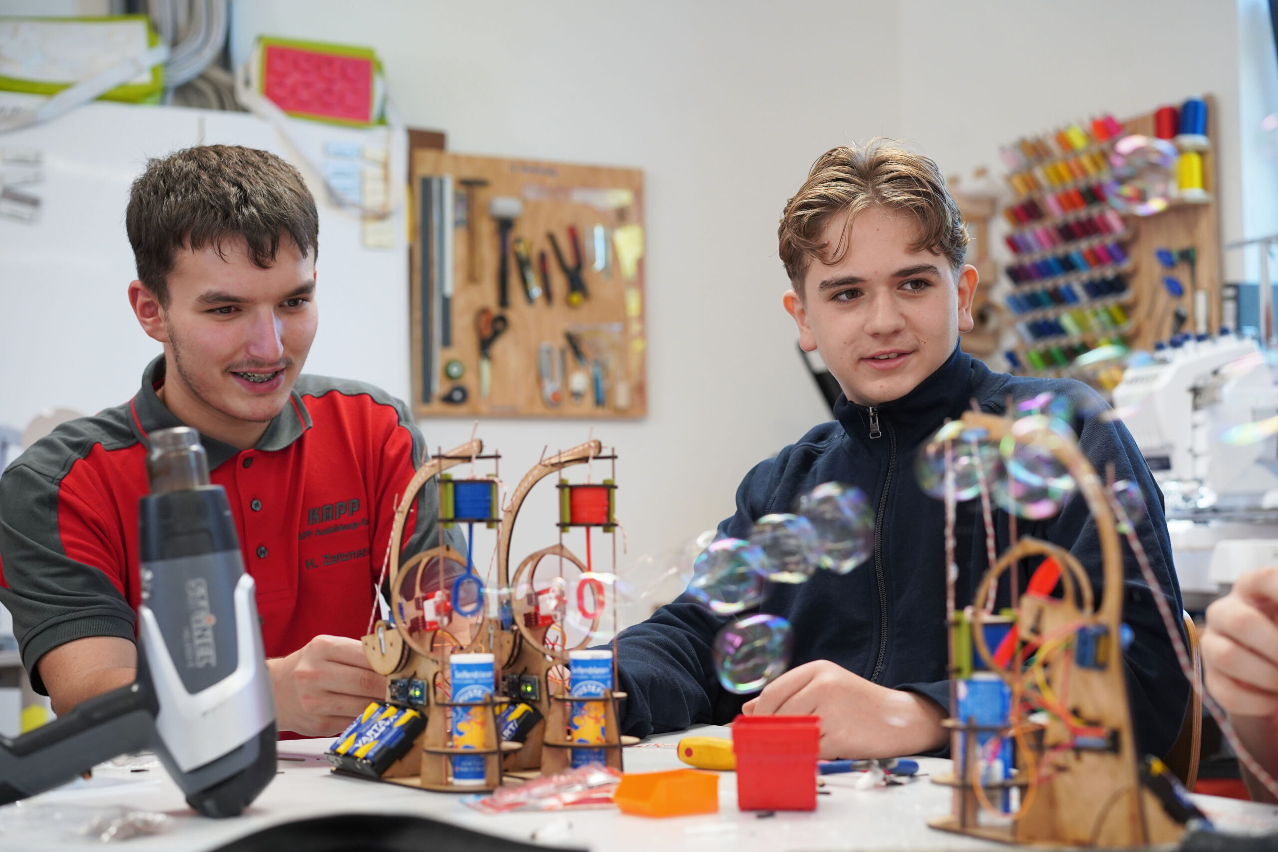 Zwei junge Männer sitzen an einem Tisch und sind in ein kreatives Projekt vertieft. Seifenblasen schweben um sie herum, verschiedene kleine Strukturen und bunte Bauteile aus ihrem Studium an der Hochschule Coburg bedecken die Oberfläche. Im Hintergrund liegen Werkzeuge, die ihre konzentrierte Hingabe unterstreichen.