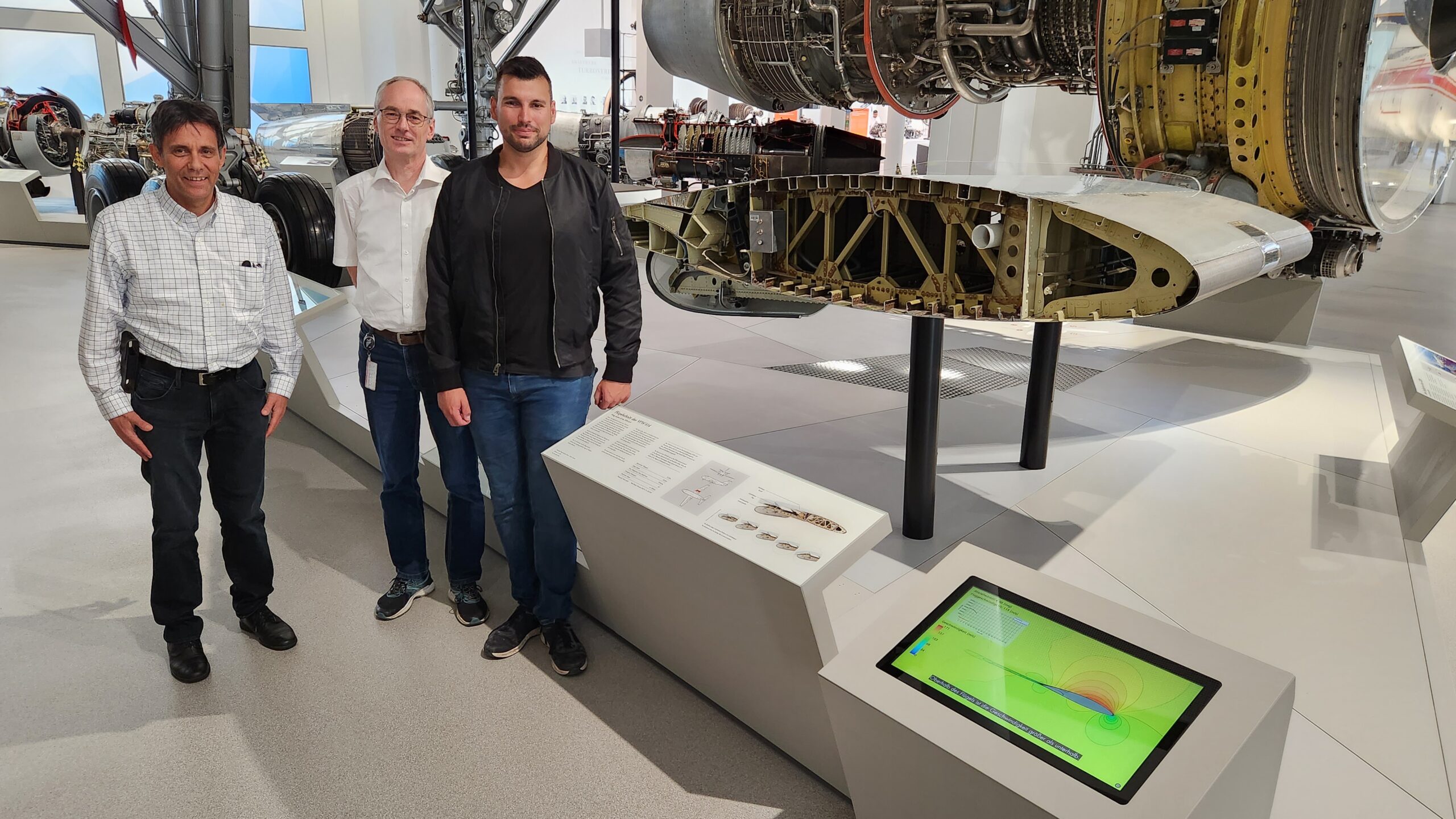 Three men stand next to a display of jet engine components at the museum, likely on a visit from Hochschule Coburg. One wears a checked shirt, another sports a white shirt, and the third dons a dark jacket. A touchscreen display is visible in the foreground, offering interactive insights.