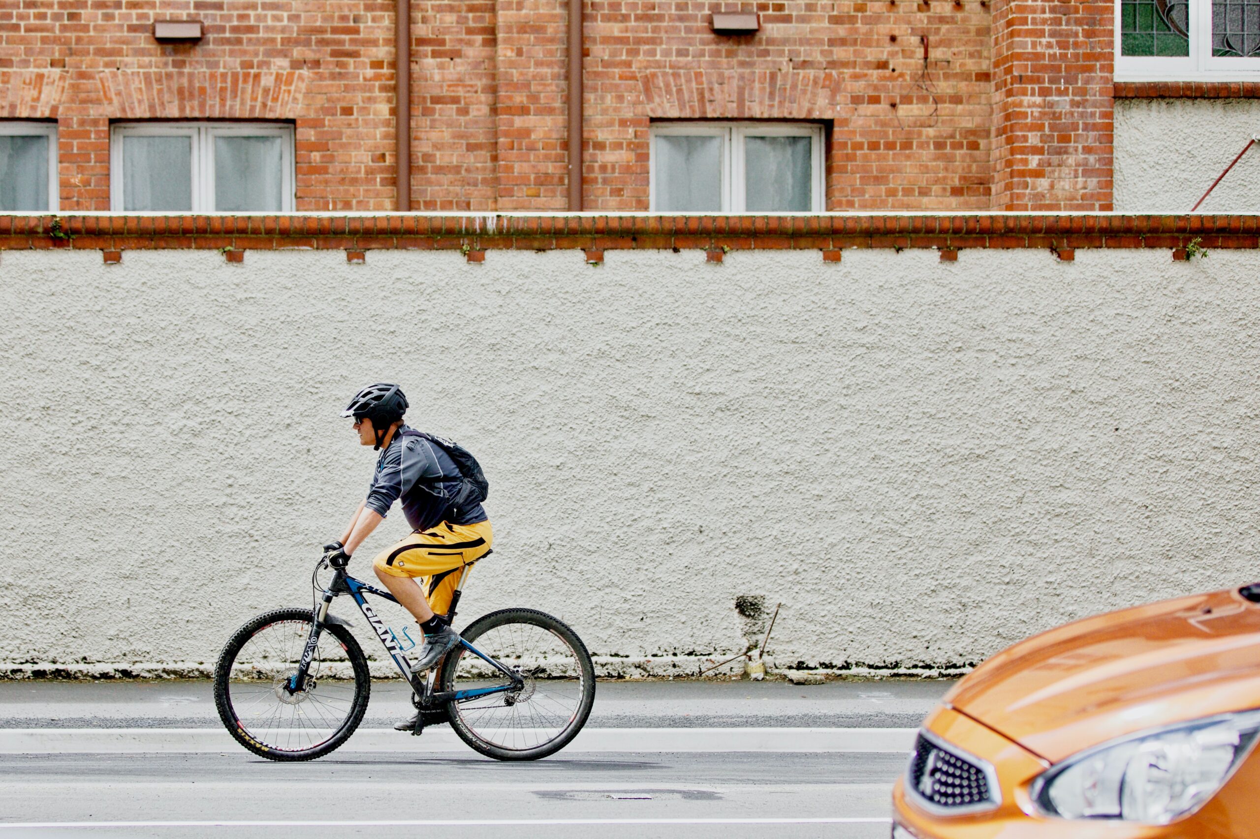 Eine Person mit Helm und Rucksack fährt mit dem Fahrrad auf einer Stadtstraße in der Nähe der Hochschule Coburg. Sie fahren an einer strukturierten weißen Wand vorbei, über der sich rote Backsteingebäude befinden, während im Vordergrund ein verschwommenes orangefarbenes Auto vorbeirast.