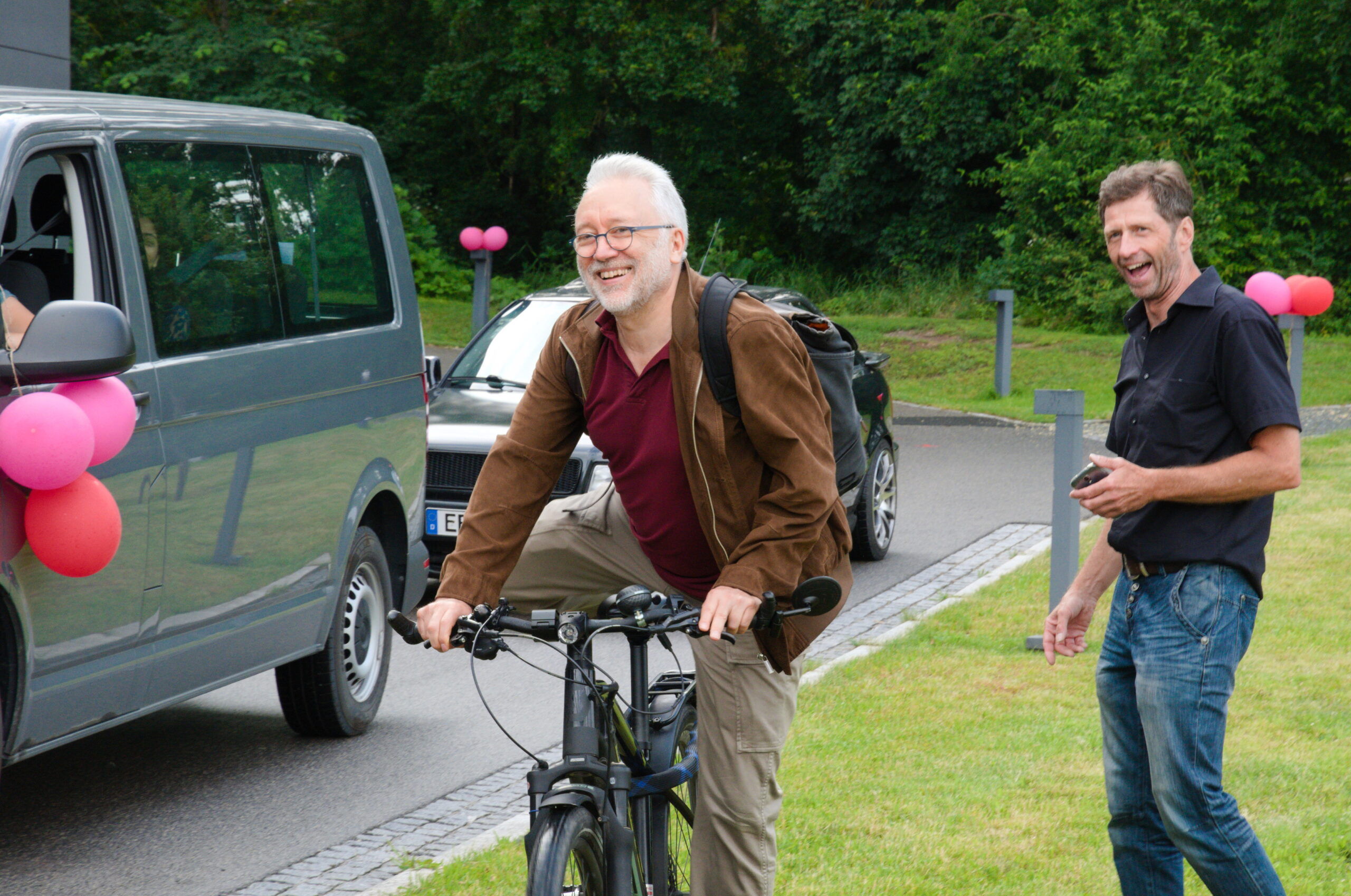 Ein Mann mit weißem Bart fährt auf seinem Fahrrad neben einem mit rosa Luftballons geschmückten Lieferwagen in der Nähe der Hochschule Coburg. Neben ihm läuft ein anderer Mann, lächelnd und mit einem Telefon in der Hand, während sie eine von Gras und Bäumen gesäumte Straße entlangfahren.