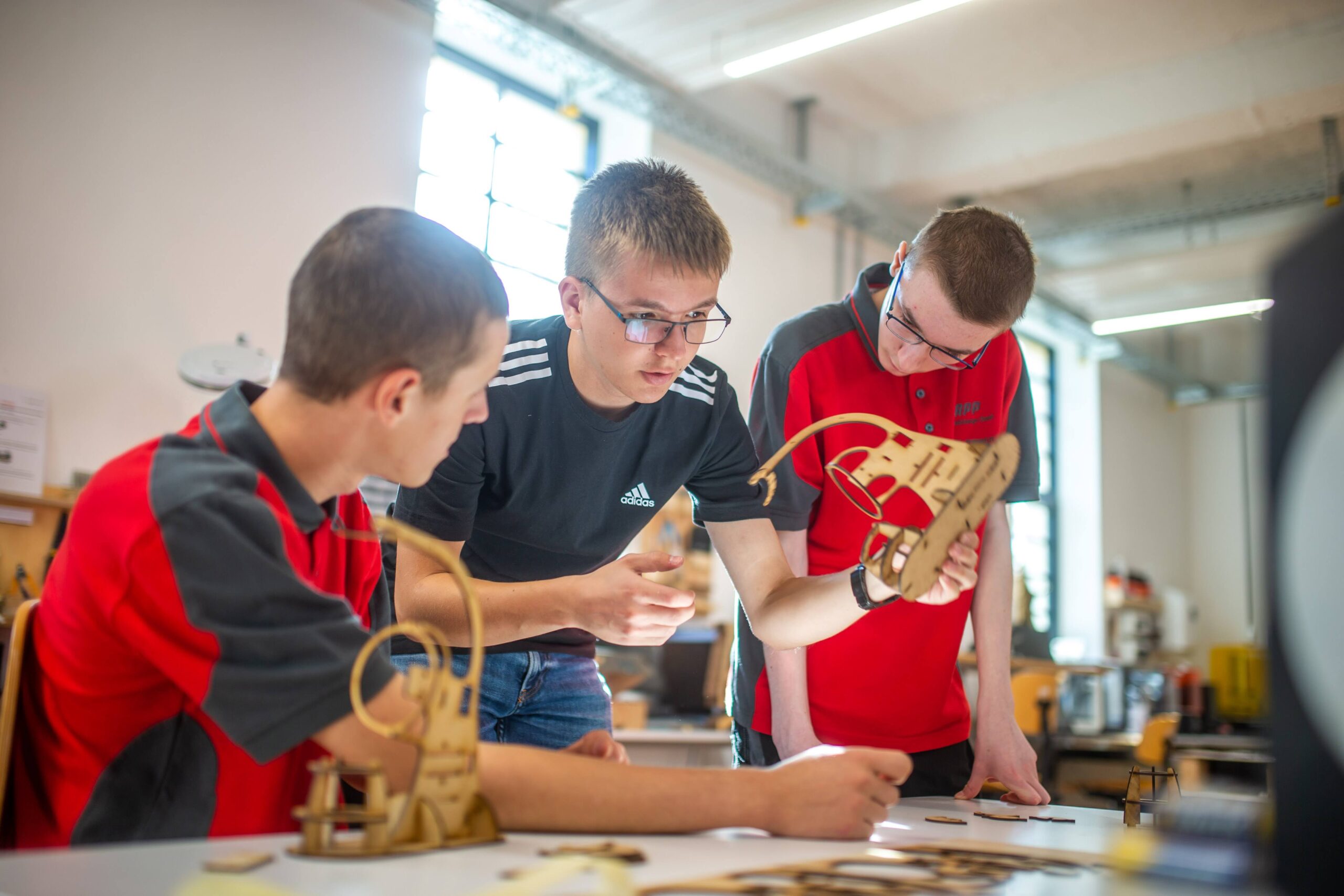 Drei junge Leute bauen an einem Tisch in einem hellen Raum der Hochschule Coburg Holzmodelle zusammen. Sie sind konzentriert und engagiert und diskutieren begeistert über ihre Arbeit. Durch ein großes Fenster im Hintergrund fällt Tageslicht ein und taucht ihr kreatives Werk in einen warmen Schein.