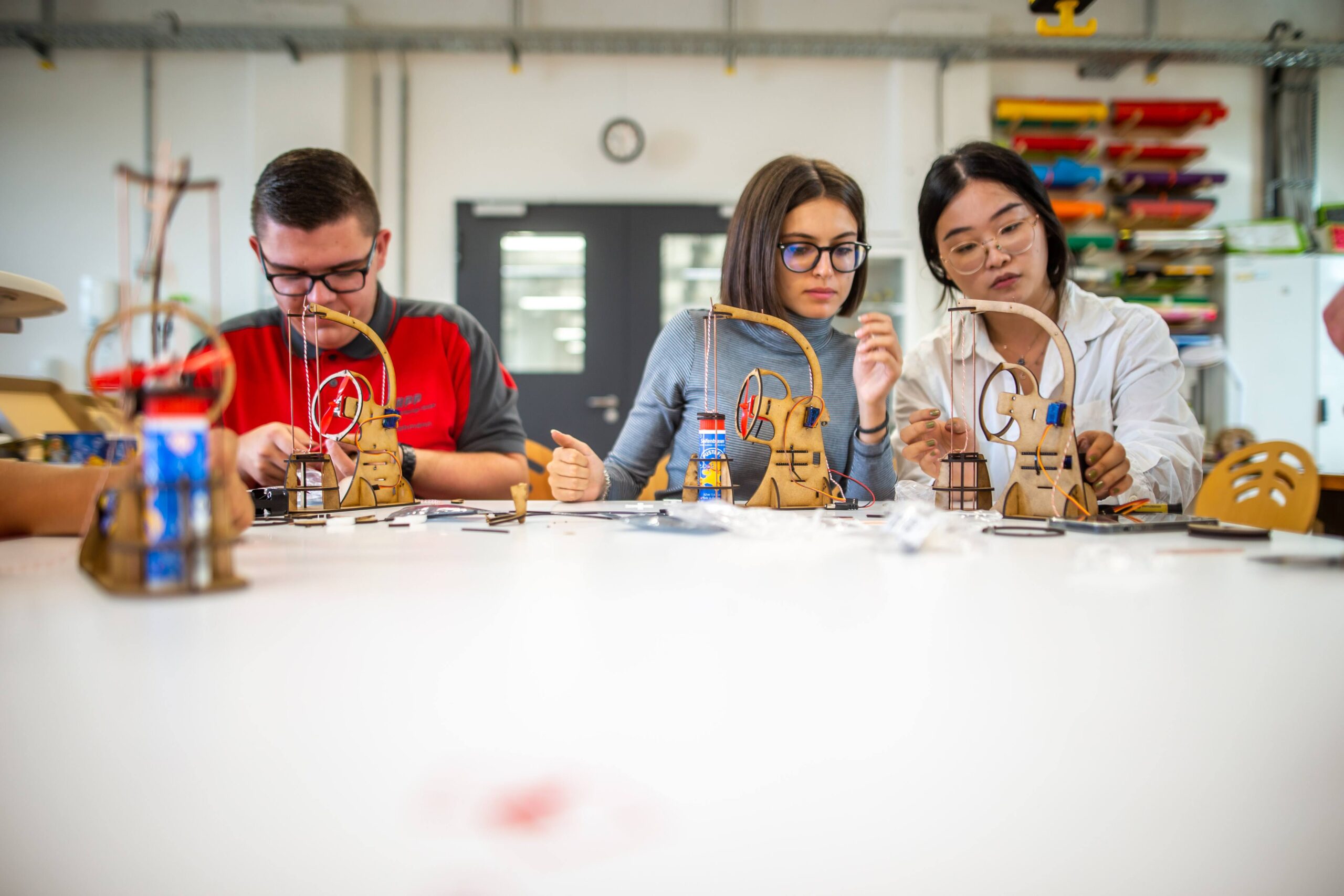 Drei Personen der Hochschule Coburg bauen konzentriert an einem Tisch in einer Werkstatt kleine Holzmodelle zusammen. Um sie herum sind verschiedene Werkzeuge und Materialien, im Hintergrund bunte Regale. Die Atmosphäre ist aufmerksam und gemeinschaftlich.