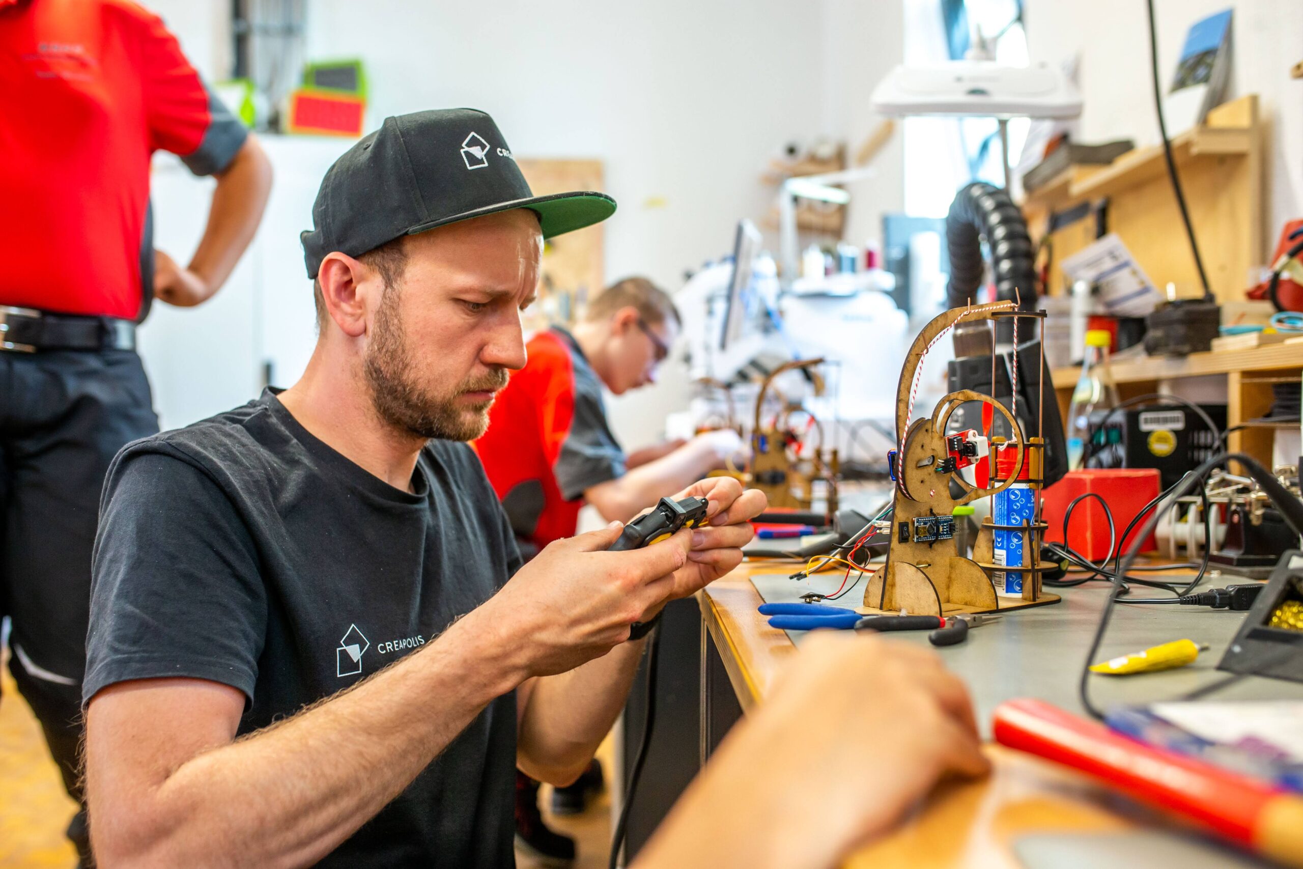 Ein Mann mit Mütze lötet konzentriert ein kleines elektronisches Bauteil an einer Werkbank voller Werkzeuge und Platinen und steht damit stellvertretend für den Innovationsgeist der Hochschule Coburg. Im Hintergrund arbeitet eine weitere Person in einer gut beleuchteten Werkstattumgebung.