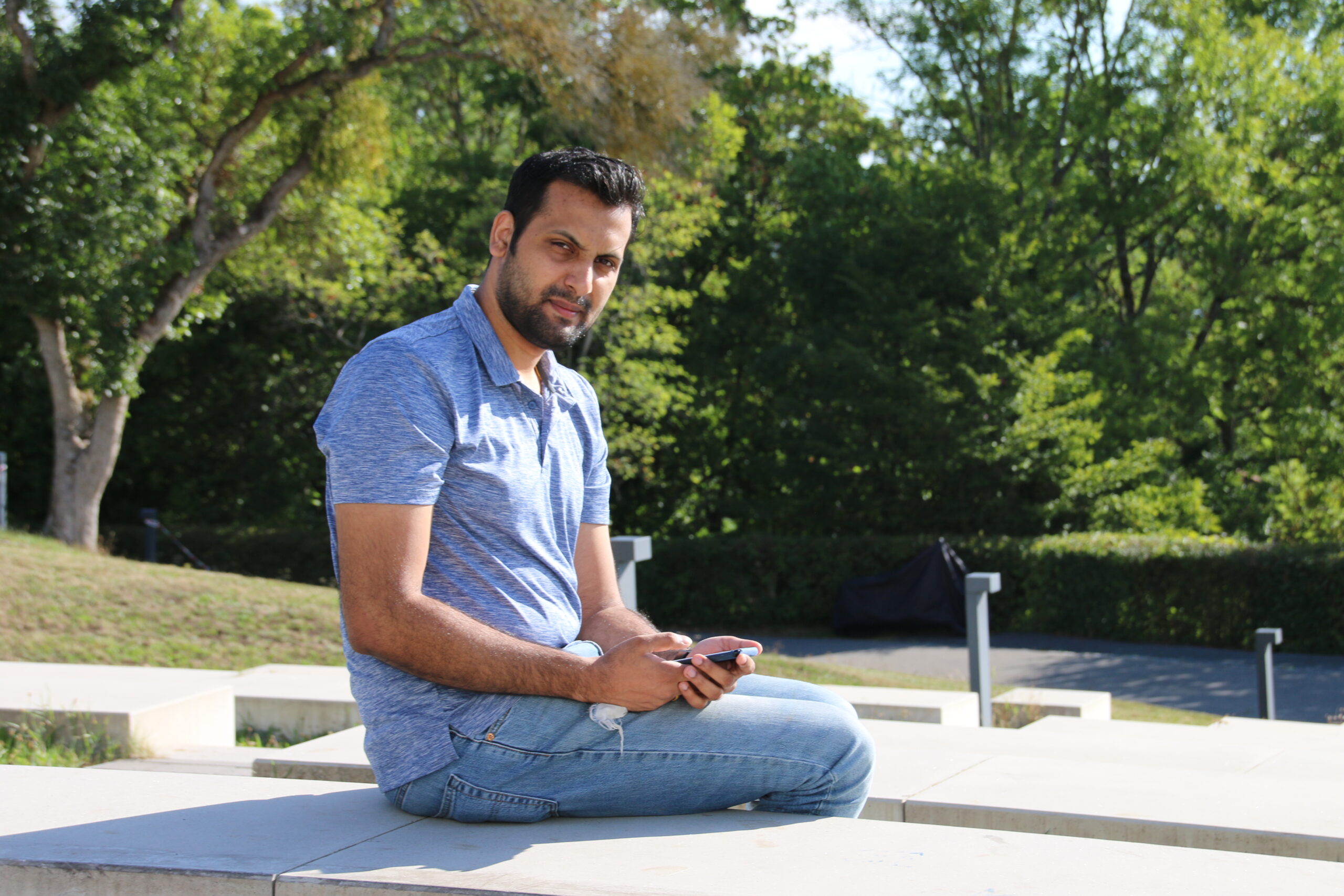 Ein Mann in blauem Hemd und Jeans sitzt draußen auf einer Steinbank an der Hochschule Coburg. Er hält ein Telefon in der Hand und blickt in die Kamera, eingerahmt von grünen Bäumen und Sonnenlicht.