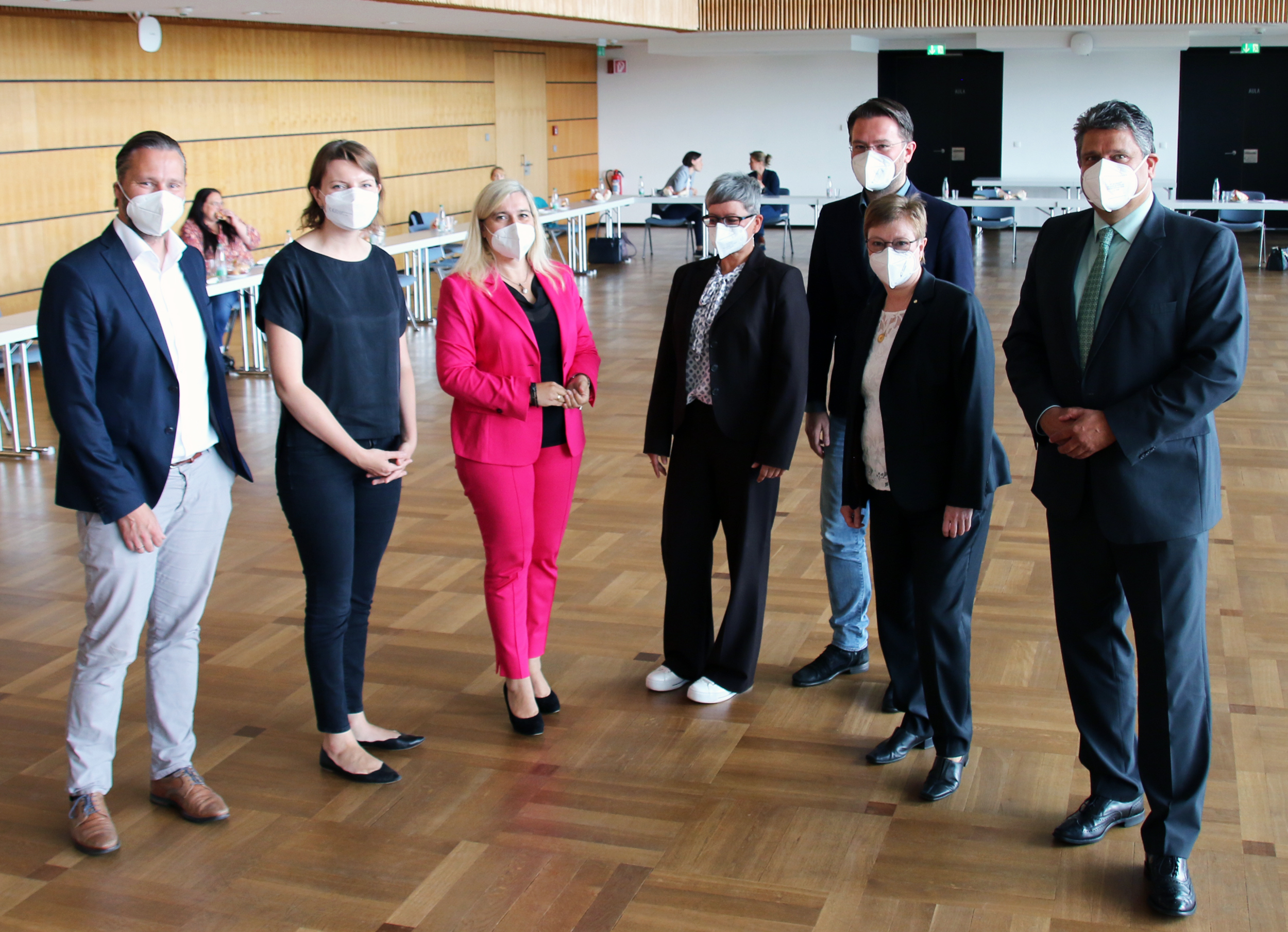 Sieben Personen stehen im Innenbereich der Hochschule Coburg, alle tragen Gesichtsmasken. Sie tragen eine Mischung aus formeller und halbformeller Kleidung. Einige andere sitzen an Tischen im Hintergrund, umgeben von dem Holzboden und den Holzwänden des Raums.