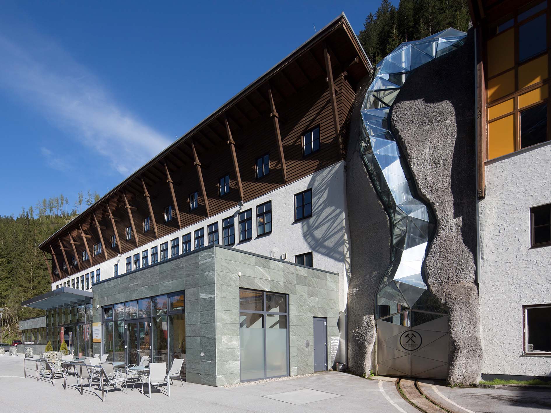 Modernes Gebäude mit Holz- und Glaselementen, an dessen Fassade sich eine abstrakte Glas- und Metallskulptur befindet. Vor einem klaren blauen Himmel und umgeben von Bäumen befindet sich dieses architektonische Juwel in der Nähe der Hochschule Coburg. Im Vordergrund befinden sich einladende Sitzbereiche im Freien.