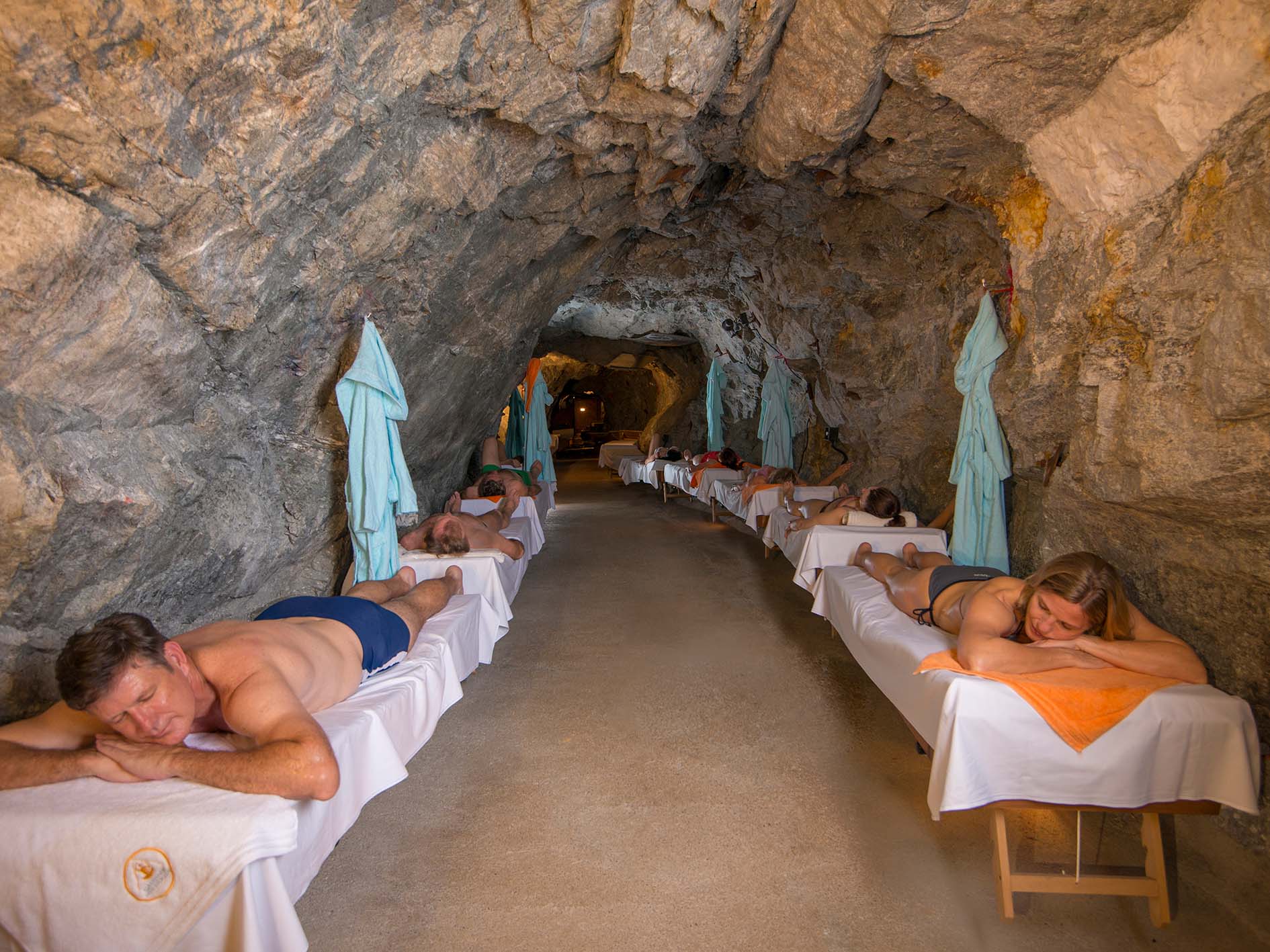 In einer Felsenhöhle, die an eine Kunstinstallation der Hochschule Coburg erinnert, liegen Menschen auf Massagetischen, die mit weißen Laken bedeckt sind. Die Höhlenwände sind aus Felsgestein, an den Wänden hängen blaue Handtücher. Das Umgebungslicht ist warm und schafft eine entspannende Atmosphäre.