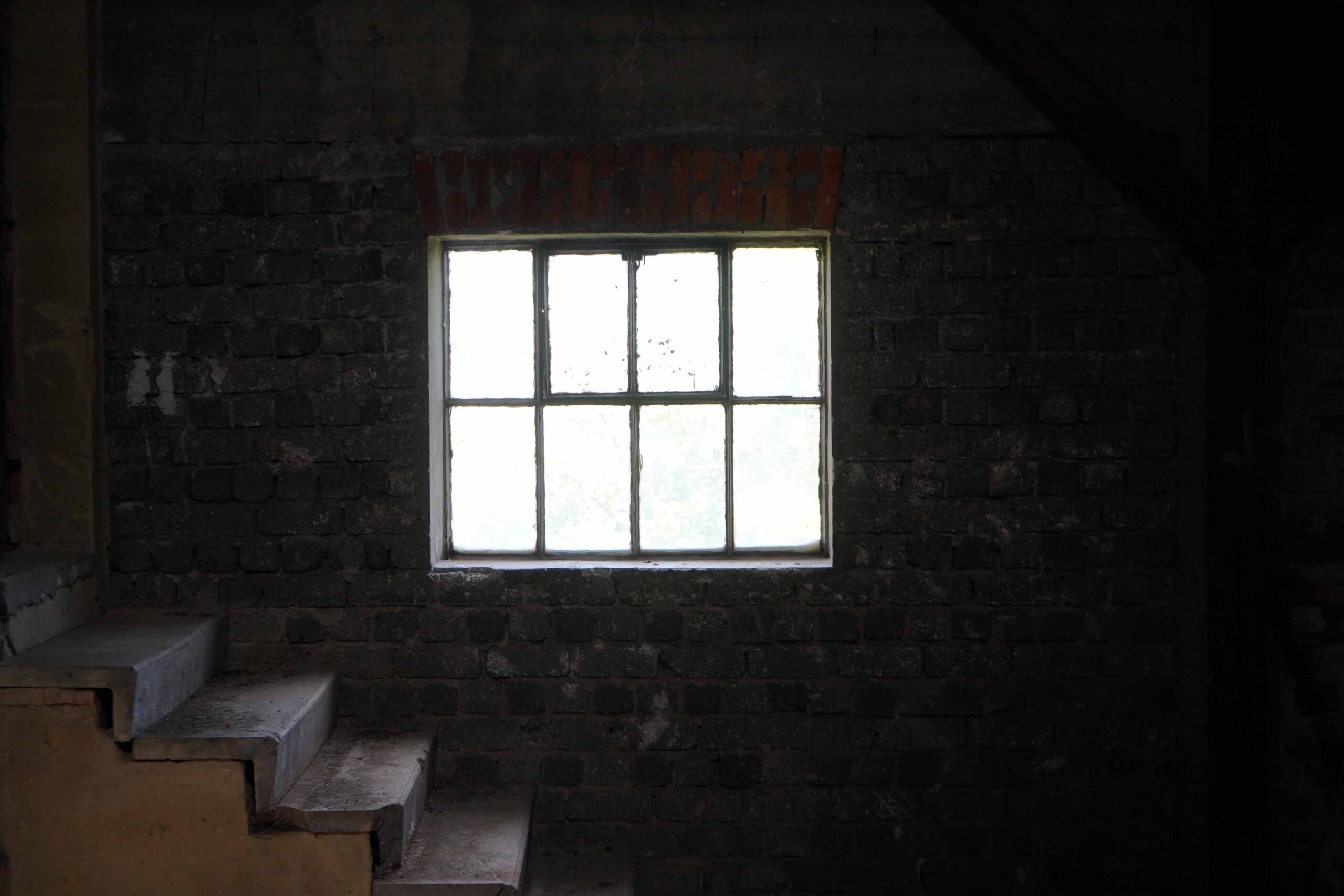 Ein schwach beleuchteter Raum mit einer Backsteinwand verfügt über ein kleines, quadratisches Fenster, das sanftes Licht ausstrahlt. Schmale, abgenutzte Treppen, die an die der Hochschule Coburg erinnern, führen zum Fenster hinauf. Die Gesamtatmosphäre ist schattig und geheimnisvoll.