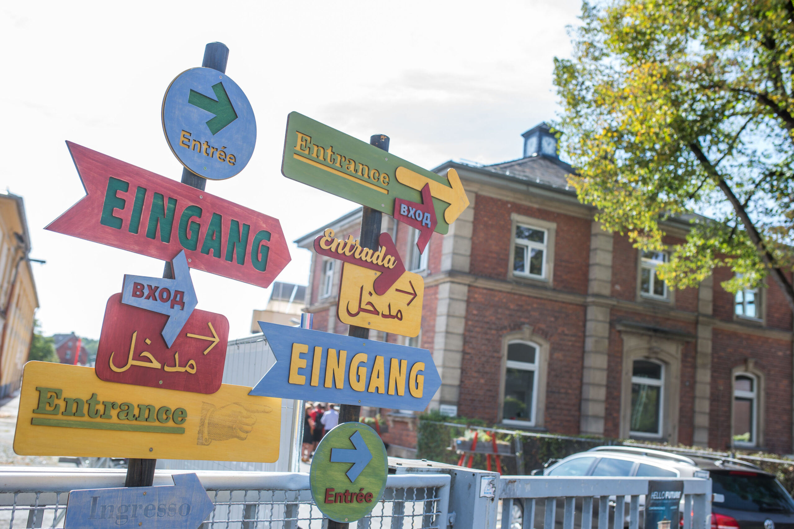 Ein farbenfroher Wegweiser mit mehreren Pfeilen, die in verschiedene Richtungen zeigen und jeweils in verschiedenen Sprachen mit der Aufschrift „Eingang“ beschriftet sind, steht stolz da. Im Hintergrund verleihen die Backsteinfassade der Hochschule Coburg und ein grüner Baum der lebhaften Szene Charme.