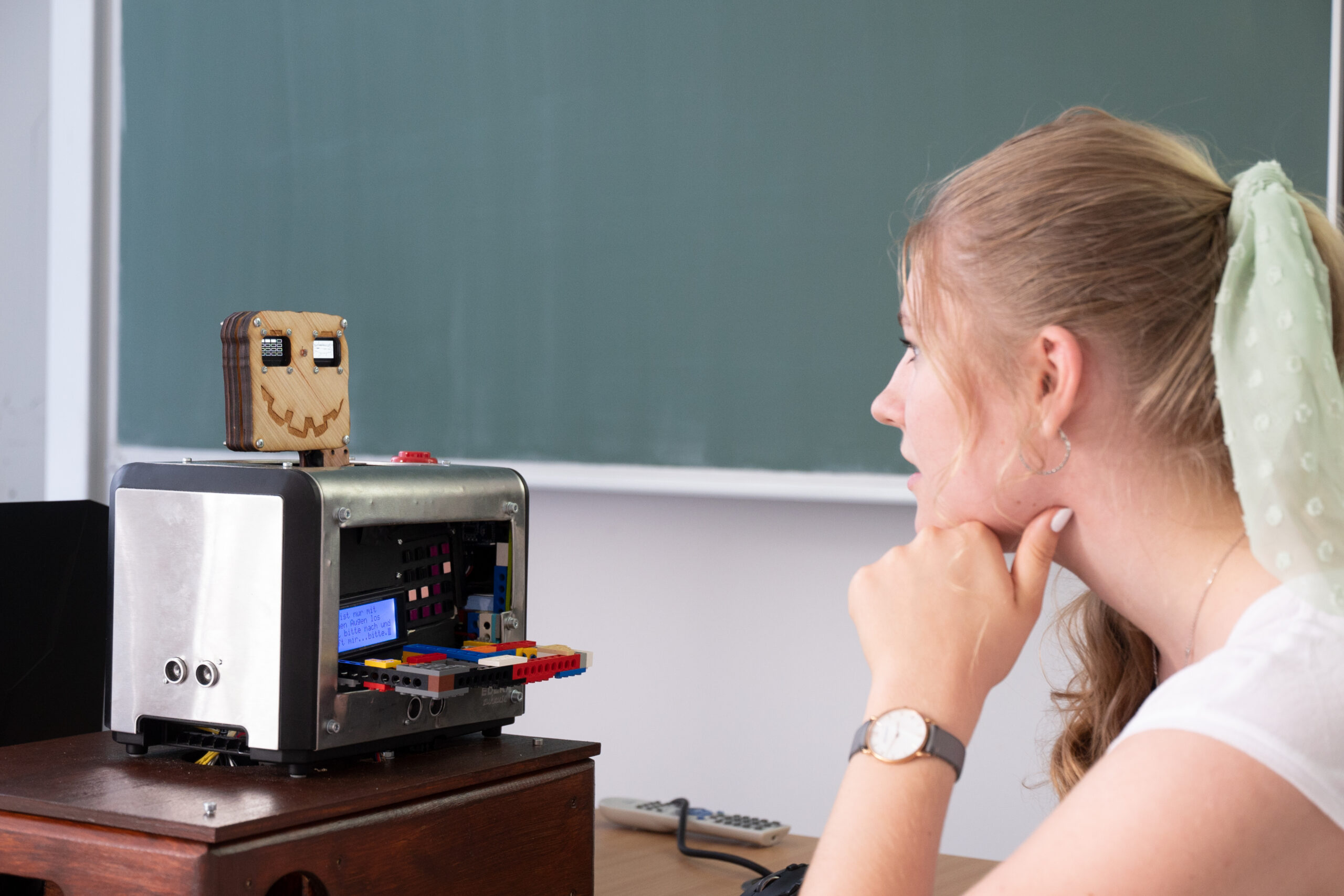 An der Hochschule Coburg sitzt eine Frau an einem Schreibtisch und blickt nachdenklich auf einen kleinen selbstgebauten Roboter mit einem Smiley auf dem Bildschirm. Der Roboter ruht auf einer Holzkiste, während eine grüne Tafel den Hintergrund bildet, während sie über ihre nächste Innovation nachdenkt.