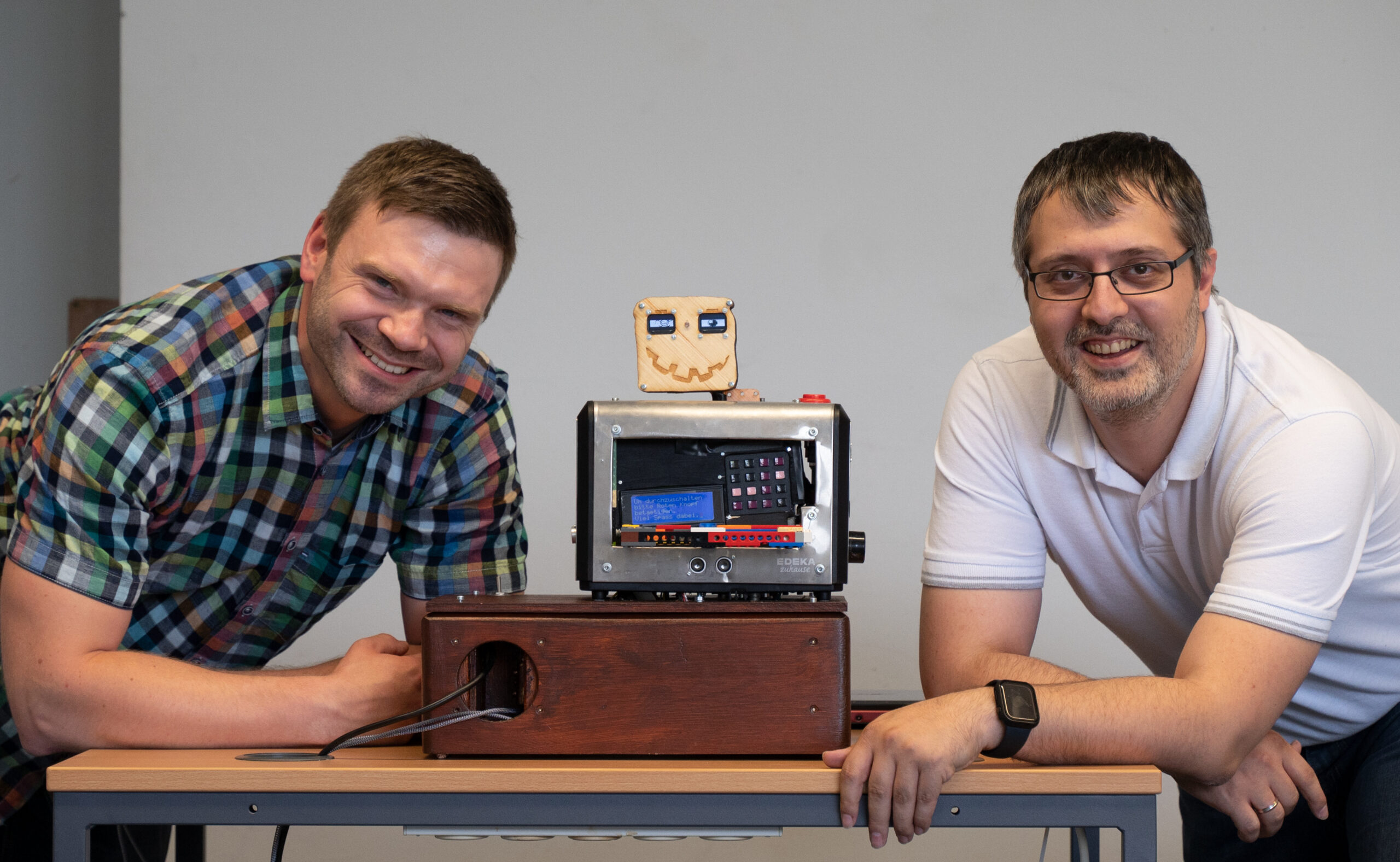 Zwei lächelnde Männer knien neben einem kleinen Roboter mit Smiley auf einer Holzkiste. Der Mann links trägt ein kariertes Hemd, während sein Begleiter im weißen Poloshirt stolz die Hochschule Coburg repräsentiert. Der Roboter präsentiert eifrig seine komplizierten elektronischen Komponenten.