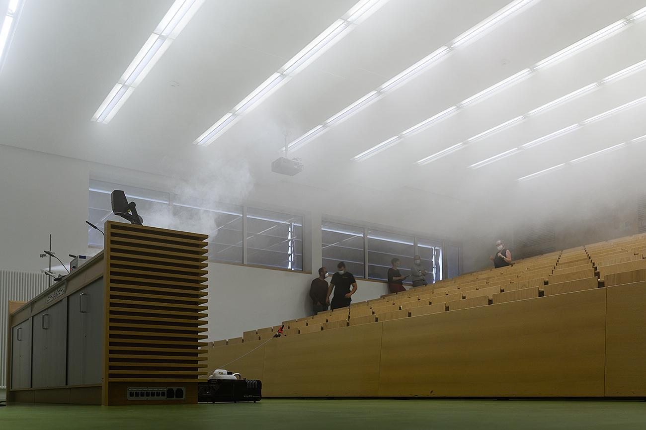 Ein Hörsaal mit Holzsitzen an der Hochschule Coburg füllt sich für eine Demonstration mit Rauch. Beobachter stehen an den Fenstern und beobachten die Szene. Der Raum ist durch Deckenlampen gut beleuchtet und an prominenter Stelle steht ein Rednerpult.