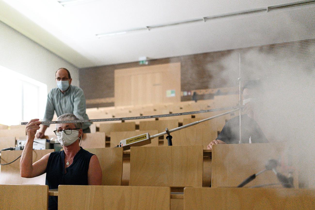 In einem Hörsaal der Hochschule Coburg führen drei Personen ein Experiment durch. Einer misst die Entfernung mit einem Maßband, ein anderer bedient die Ausrüstung und ein dritter sitzt inmitten von weißem Rauch oder Nebel. Alle tragen Masken, was darauf hindeutet, dass es sich bei dem Experiment um Luftströmungen oder Diffusion handelt.