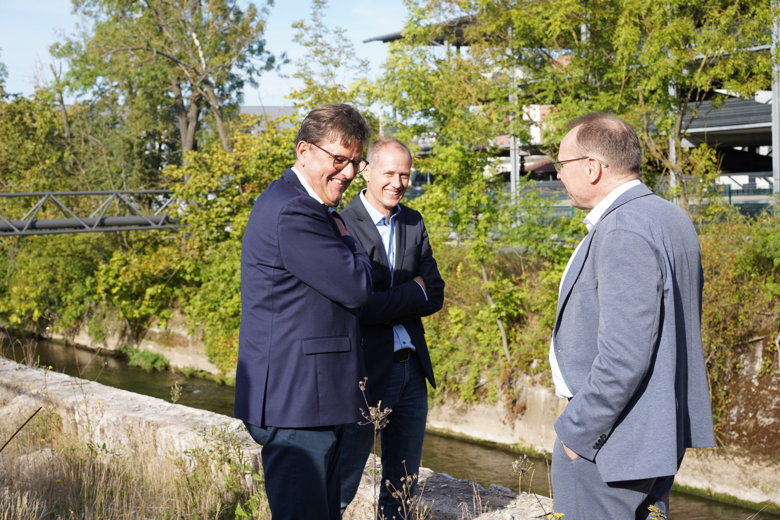 Drei Männer in Anzügen stehen im Freien neben einem Bach, unterhalten sich und lächeln. Im Hintergrund sind Bäume und die Metallbrücke der Hochschule Coburg zu sehen, die eine entspannte und ungezwungene Atmosphäre schaffen.