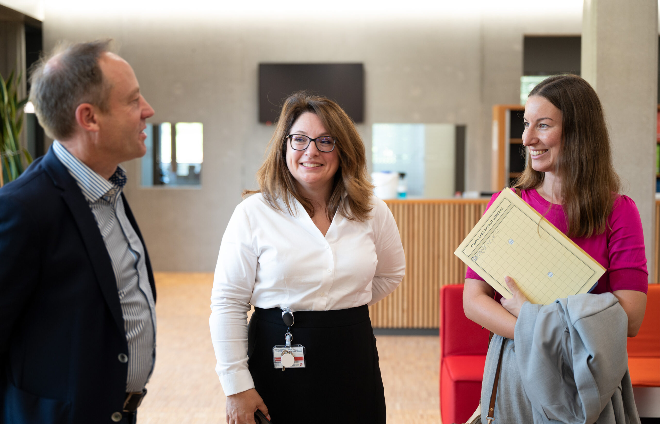 Drei Personen unterhalten sich in einem Innenbereich der Hochschule Coburg. Ein Mann im Anzug spricht mit zwei Frauen; eine trägt eine weiße Bluse und eine Brille und lächelt, die andere trägt ein rosa Oberteil und hält Dokumente und einen grauen Mantel. Der Raum ist modern eingerichtet, ein rotes Sofa ist zu sehen.