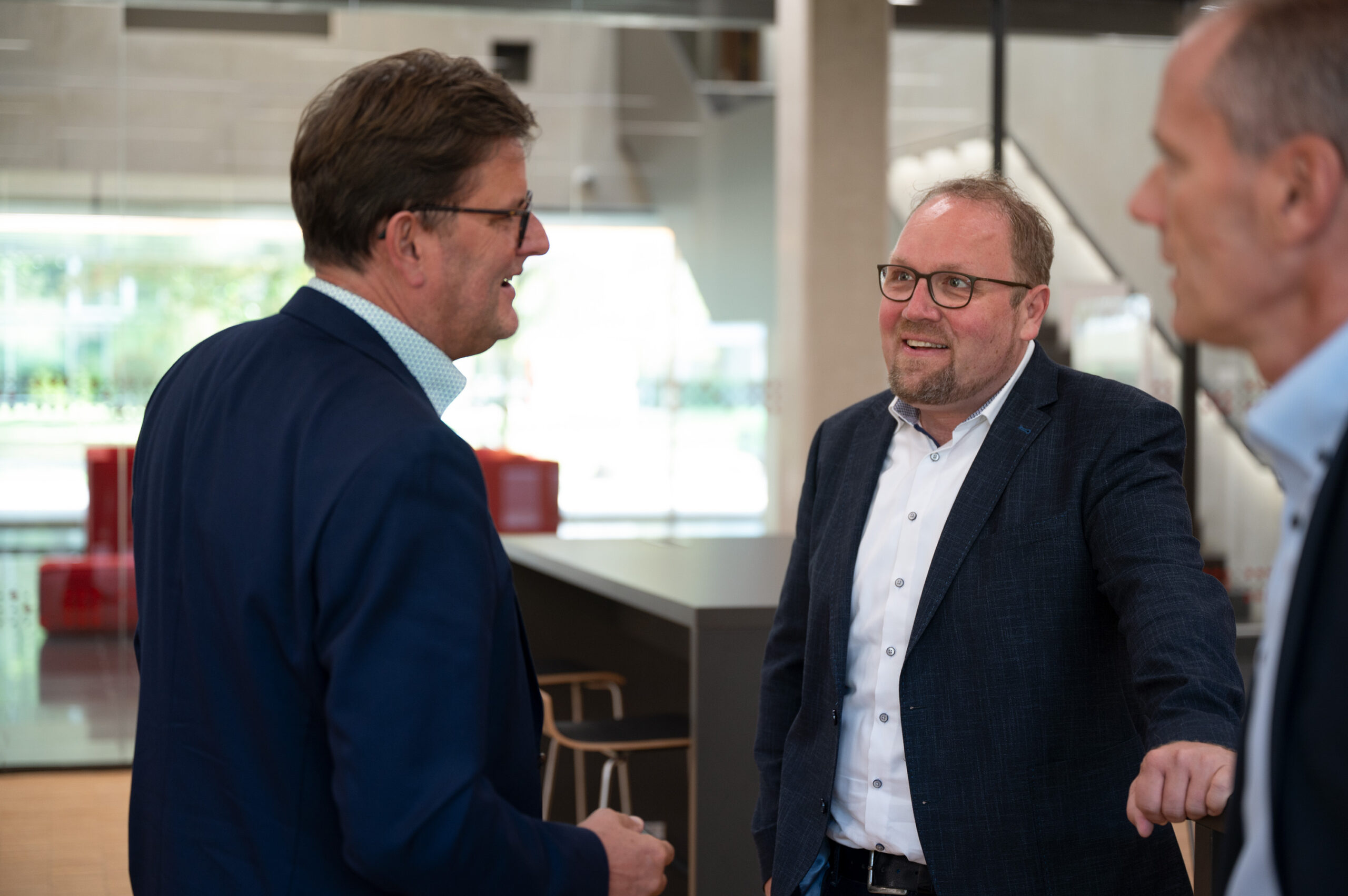 Drei Männer in Business-Kleidung unterhalten sich in einem modernen Büroraum der Hochschule Coburg mit Glaswänden. Einer spricht, die anderen hören aufmerksam zu. Im Hintergrund tragen Barhocker und Tische zur zeitgenössischen Atmosphäre bei.