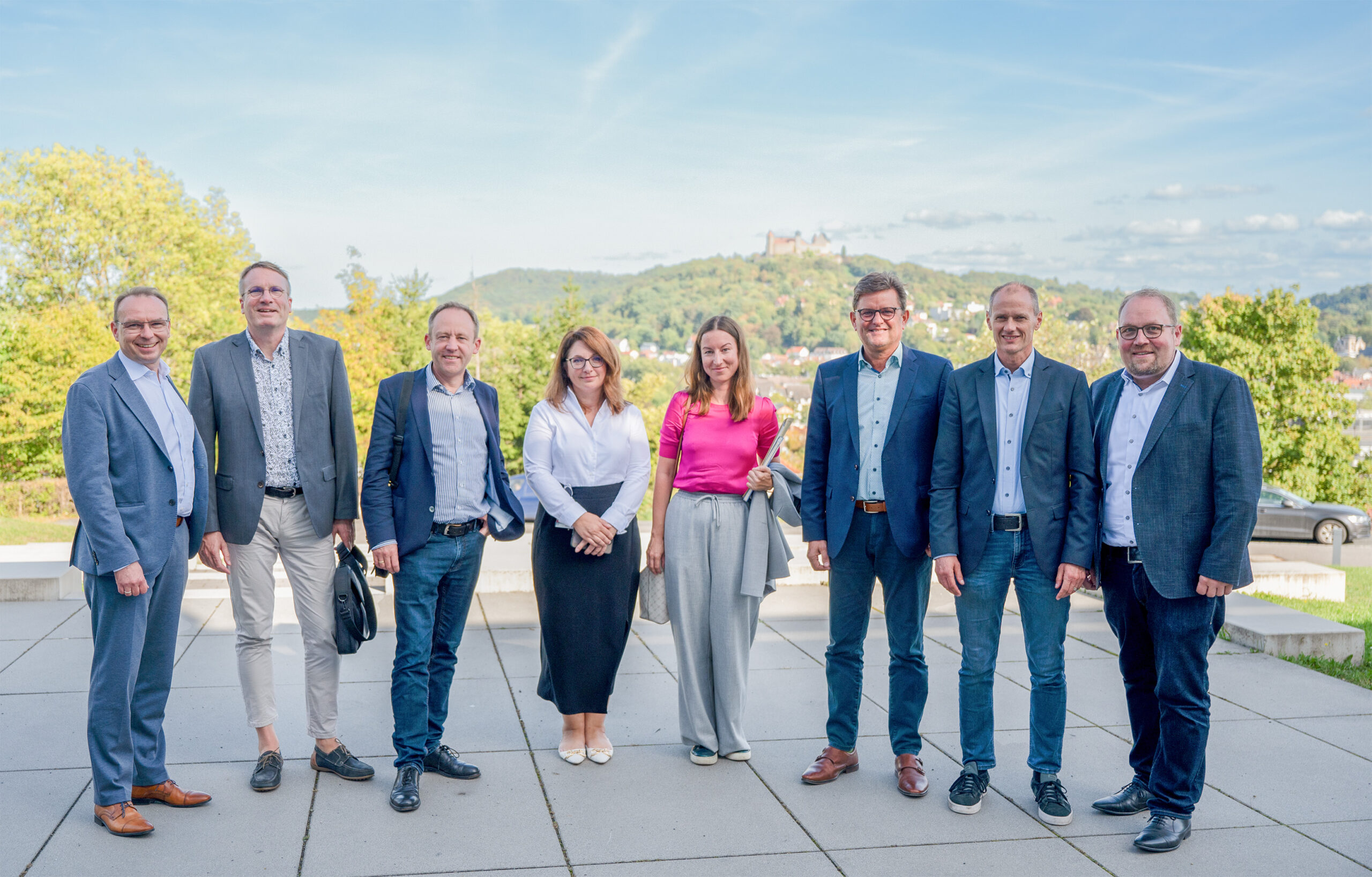 Eine Gruppe von acht Personen, fünf Männer und drei Frauen, posiert an einem sonnigen Tag im Freien an der Hochschule Coburg. Sie tragen Business-Kleidung. Hinter ihnen ist eine malerische Landschaft mit Hügeln und einem Schloss in der Ferne unter dem blauen Himmel zu sehen.