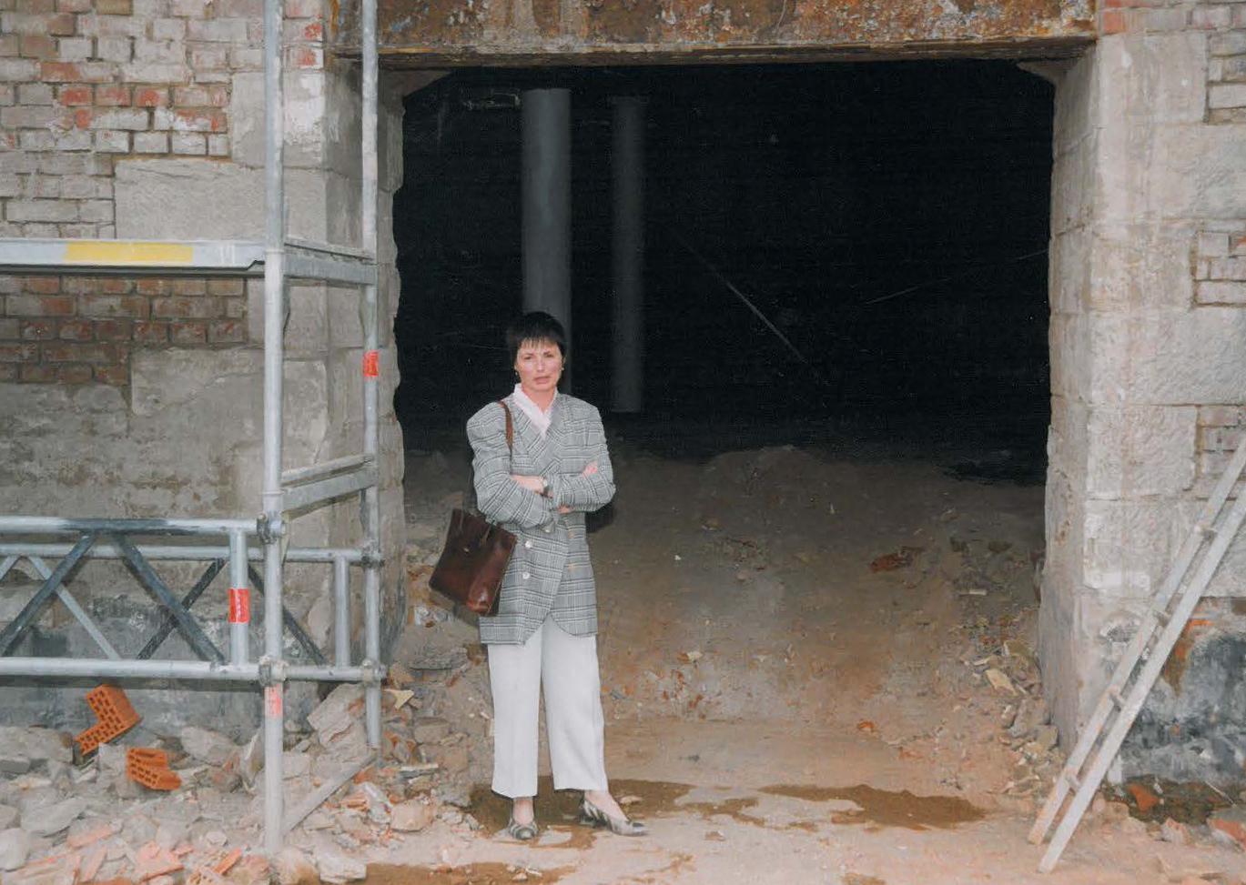 Eine Person in einer karierten Jacke steht mit verschränkten Armen auf einer Baustelle in der Nähe der Hochschule Coburg. Sie steht neben einem großen, unfertigen Eingang mit freiliegenden Ziegeln, umgeben von Gerüsten und Schutt.