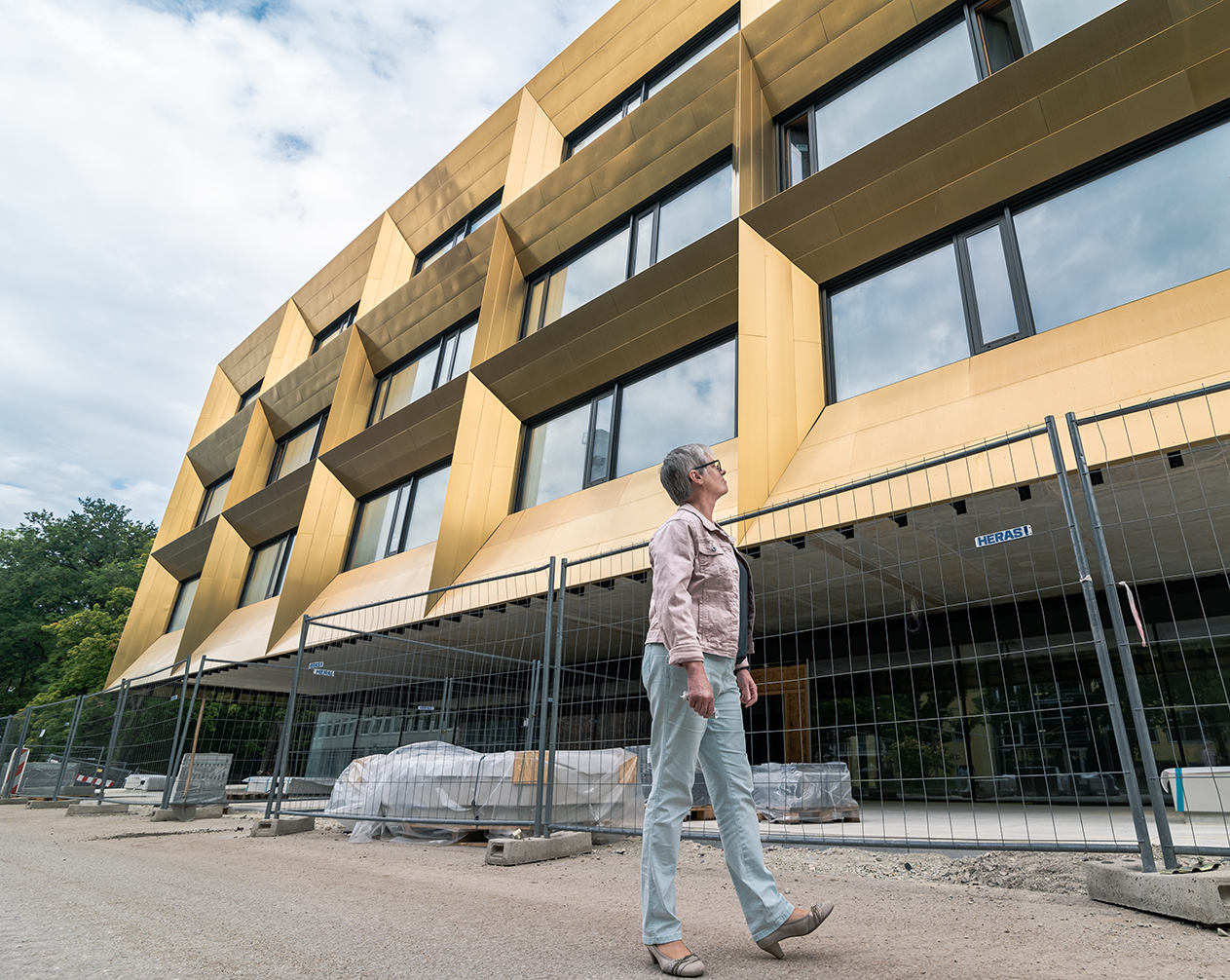 Eine Person geht am im Bau befindlichen modernen Gebäude der Hochschule Coburg vorbei, das eine einzigartige goldene Fassade aufweist. Das Gelände ist abgesperrt, Baumaterialien liegen verstreut auf dem Boden. Der bewölkte Himmel bietet eine dramatische Kulisse für dieses architektonische Unterfangen.