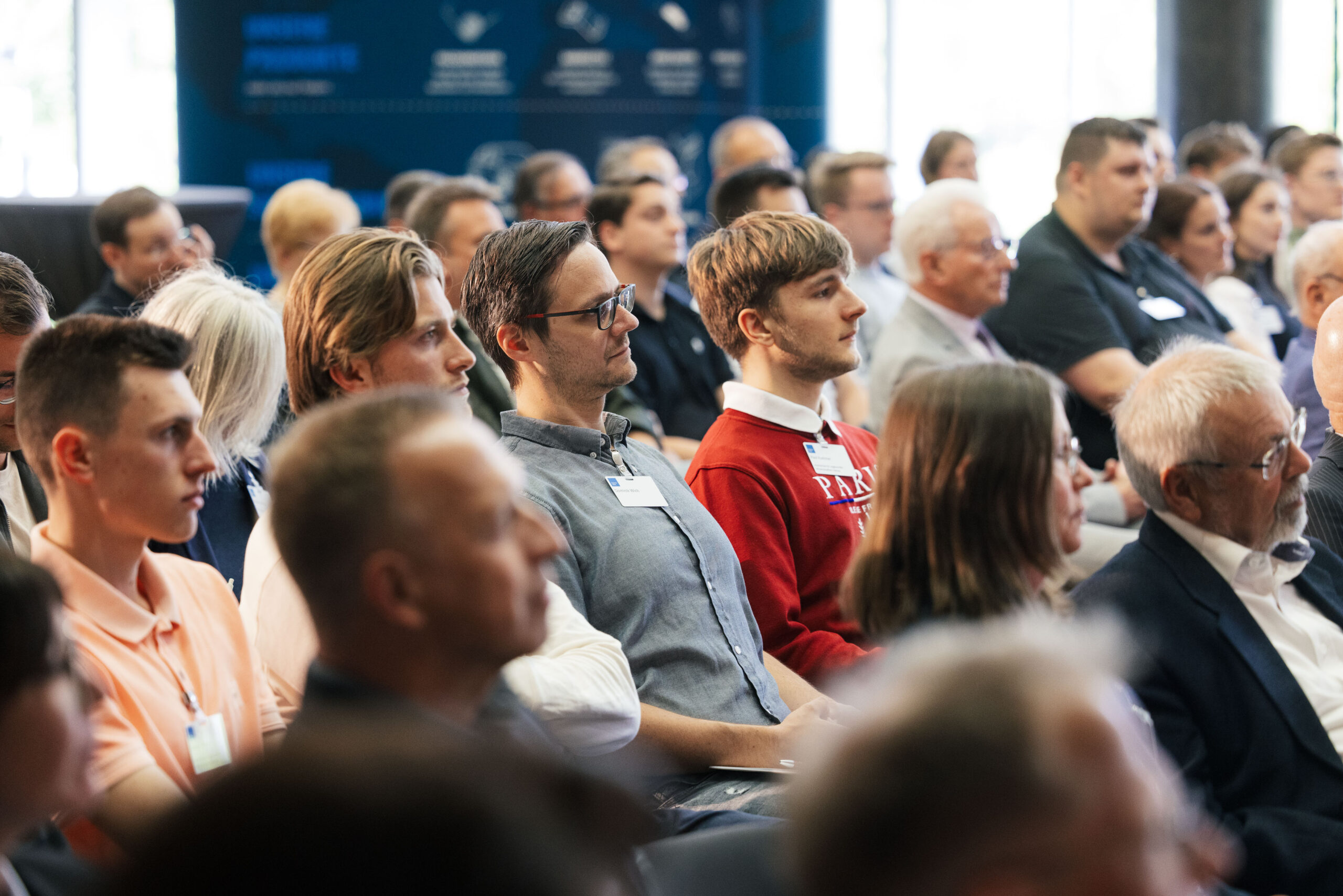 A diverse group of people seated and attentively listening at the Hochschule Coburg conference. The attendees, both men and women, are casually dressed, with some wearing name tags. A blurred background suggests a professional event setting.