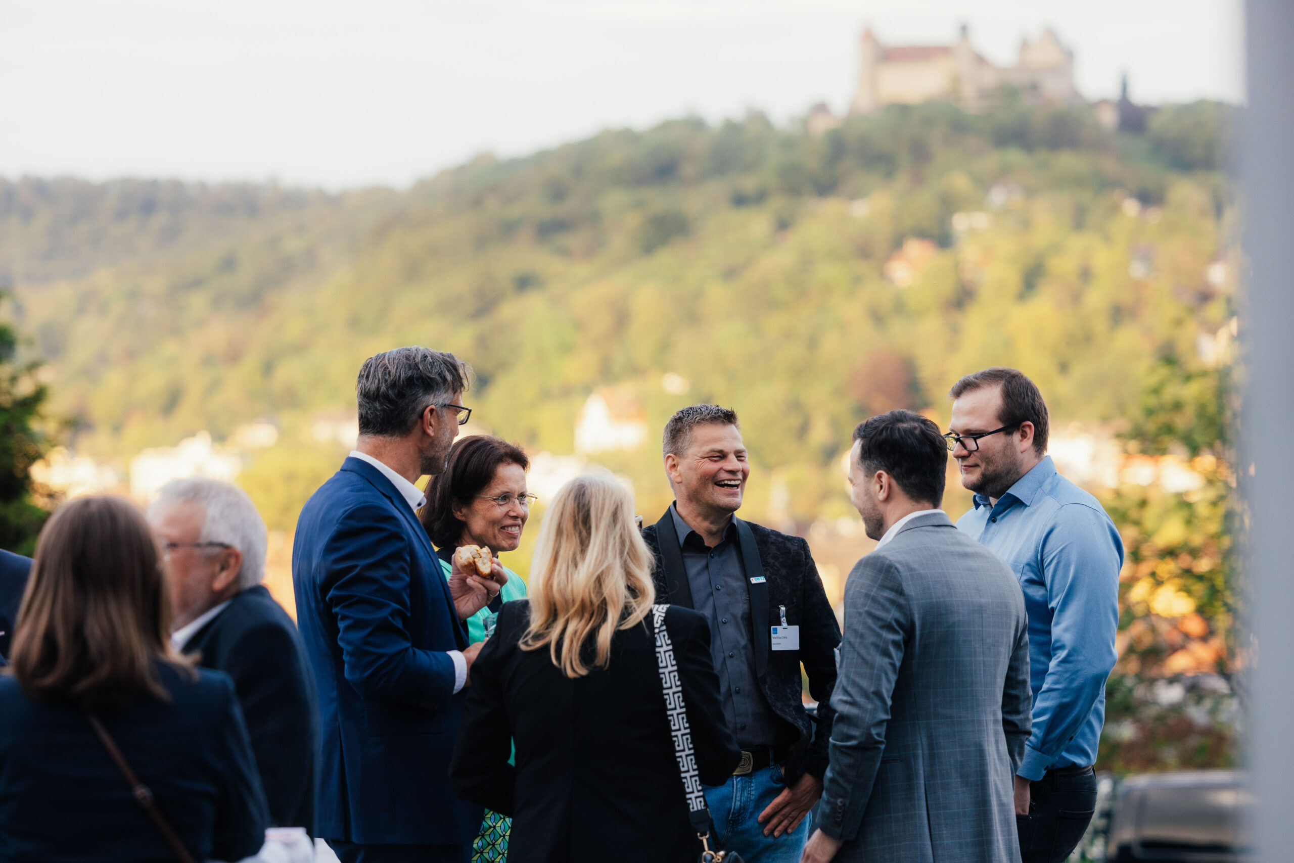 A group of people in business attire are engaged in conversation outdoors, with a scenic view of a lush hillside and the Hochschule Coburg castle in the background. The setting suggests a formal gathering or event.