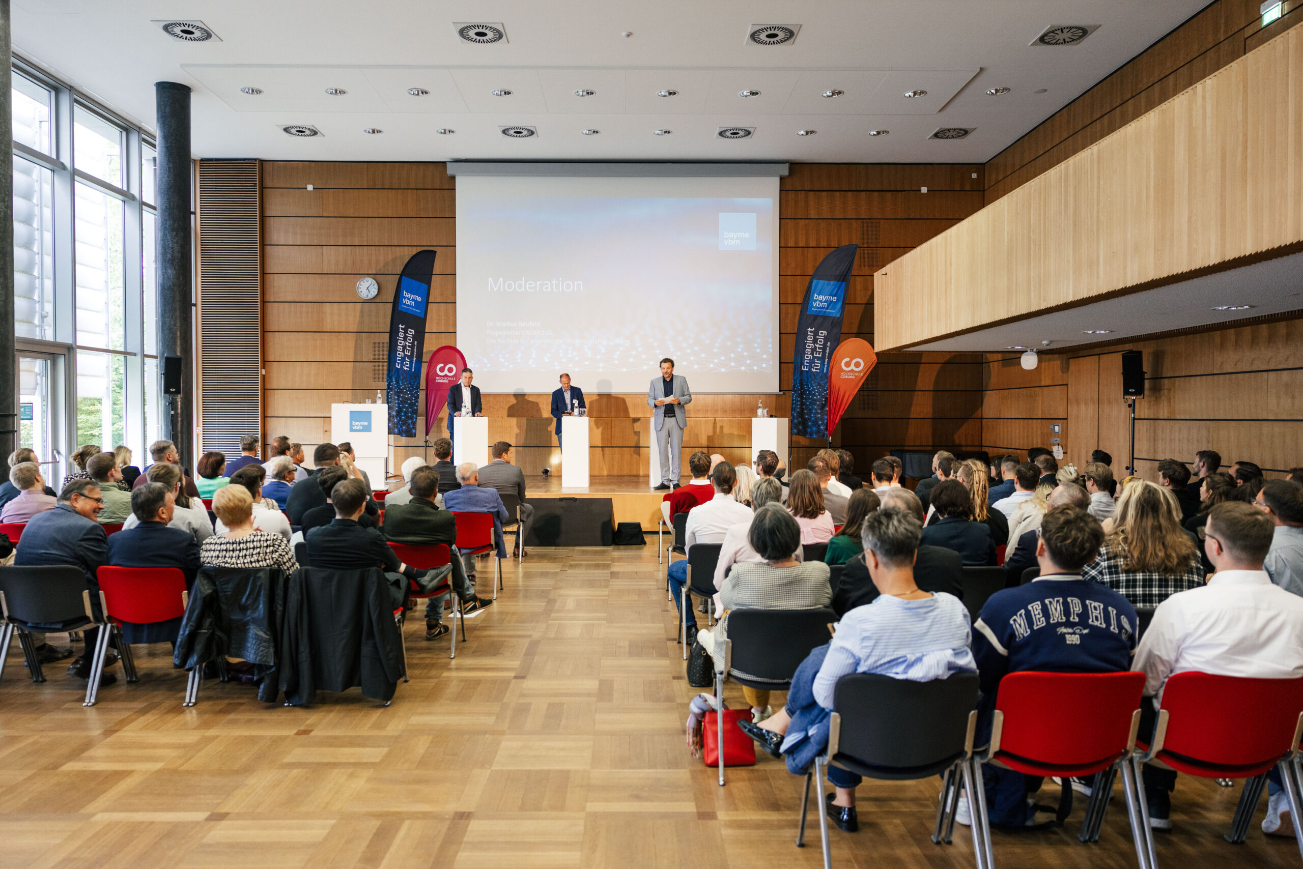 Eine große Gruppe von Menschen sitzt in Reihen und verfolgt eine Präsentation in einem modernen Konferenzraum der Hochschule Coburg. Zwei Männer stehen auf der Bühne an Rednerpulten, hinter ihnen ist eine Leinwand mit dem Wort "Moderation" zu sehen. Der Raum ist mit Holz getäfelt und hat große Fenster.