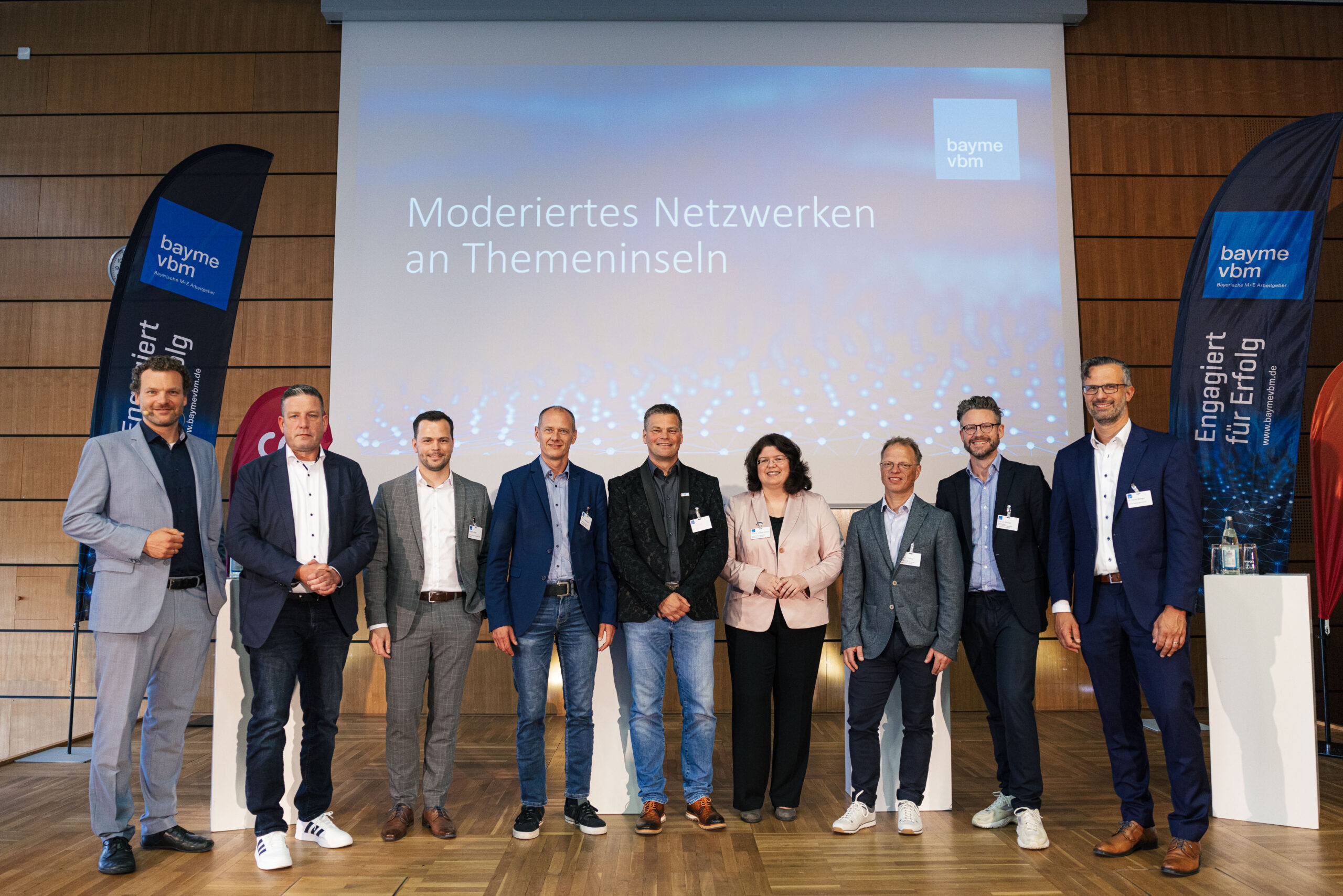 A group of nine people stands on a stage in front of a presentation screen displaying "Moderiertes Netzwerken an Themeninseln." Dressed in business attire, they are flanked by two banners with the "bayme vbm" logo. Hochschule Coburg representatives also join them to foster collaboration.