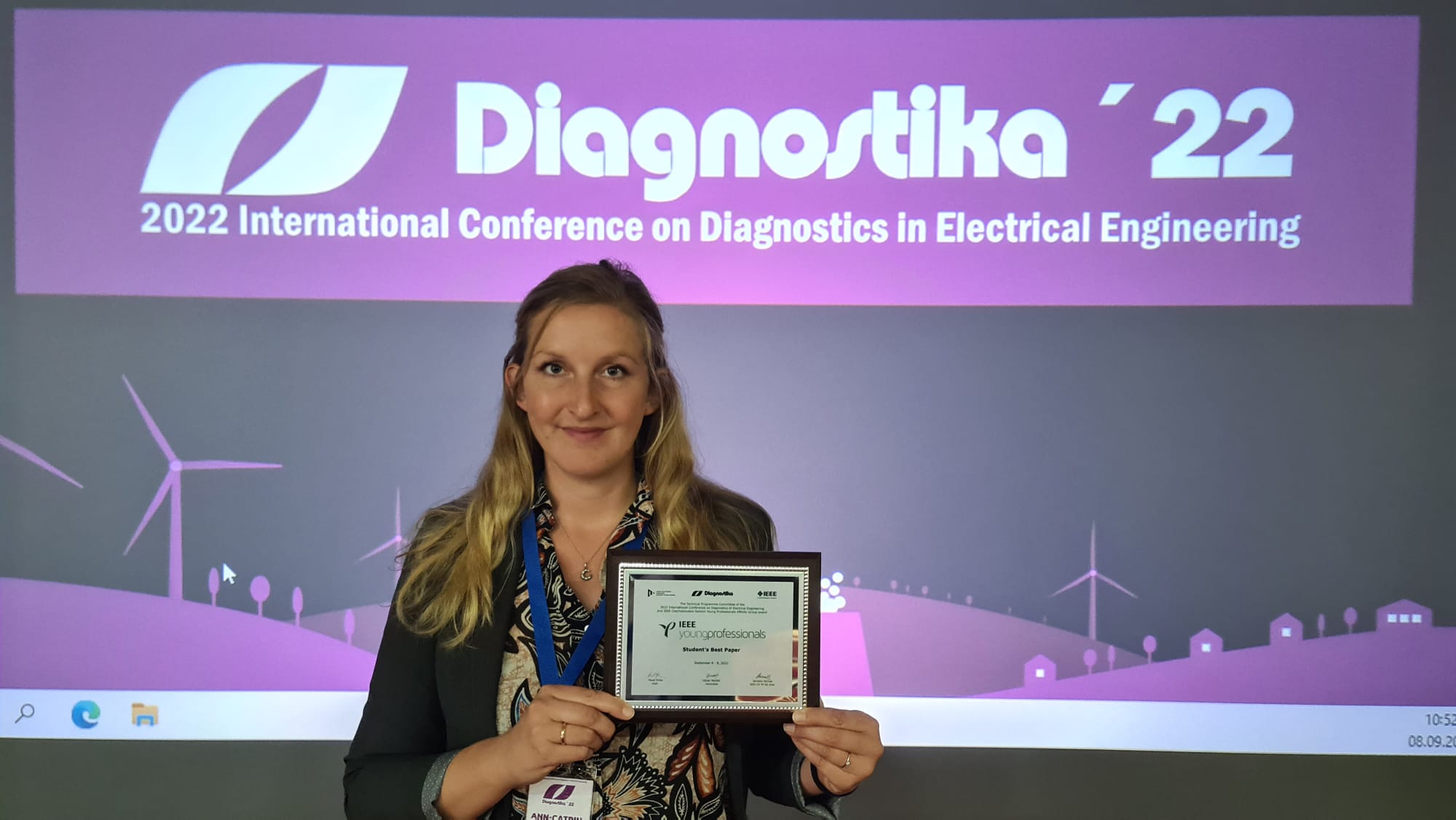 Eine Frau der Hochschule Coburg hält ein Zertifikat in der Hand und lächelt vor einem Bildschirm mit der Aufschrift „Diagnostika '22: 2022 International Conference on Diagnostics in Electrical Engineering“. Im Hintergrund sind Windräder und Gebäude in einem stilisierten Design zu sehen.