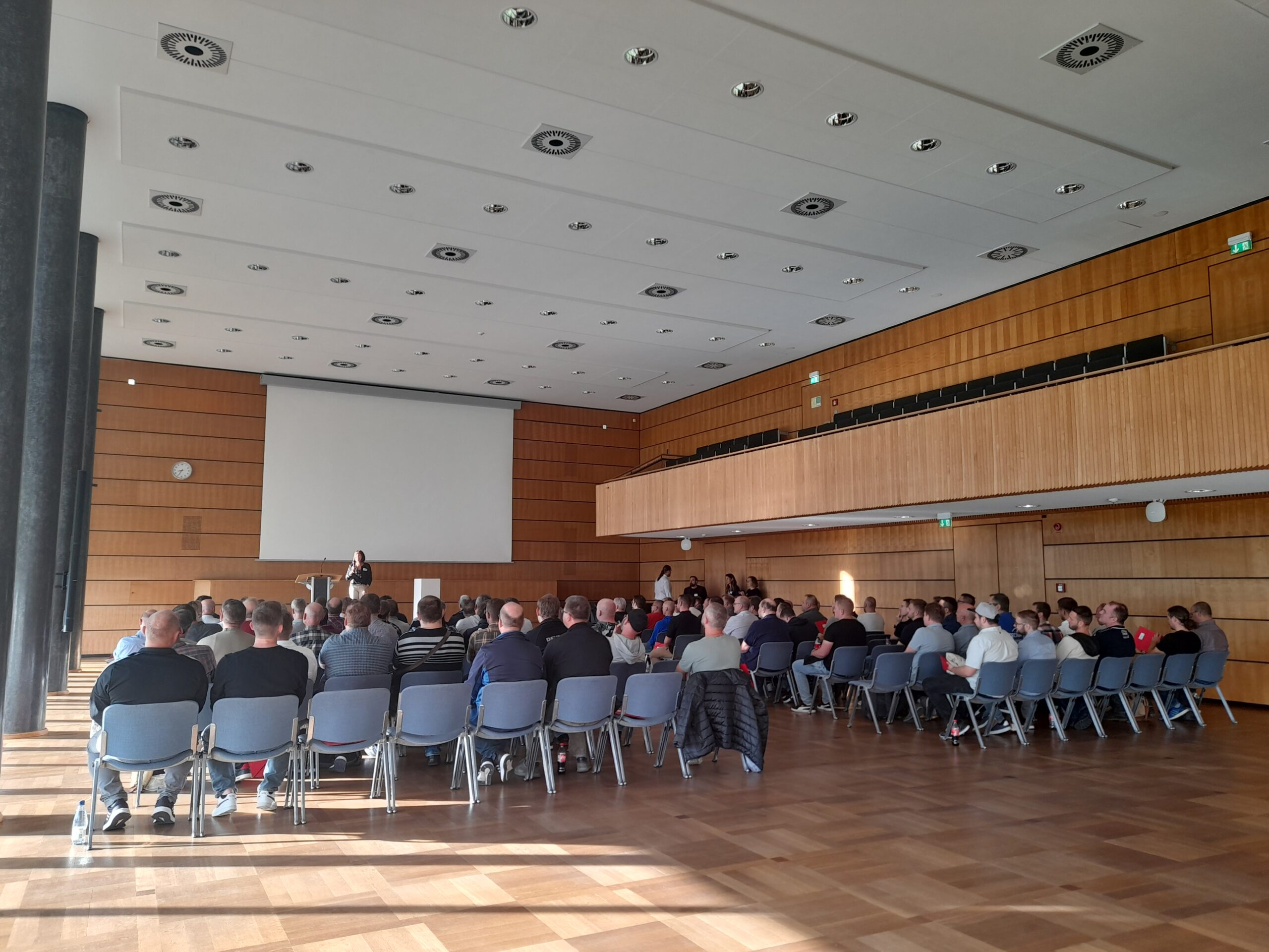 Ein geräumiger Konferenzraum an der Hochschule Coburg, in dem die Teilnehmer in Reihen einem Vortragenden am Podium gegenübersitzen. Hinter ihnen steht eine große Projektionsleinwand, während die hölzerne Innenausstattung und die großen Fenster viel natürliches Licht hereinlassen.