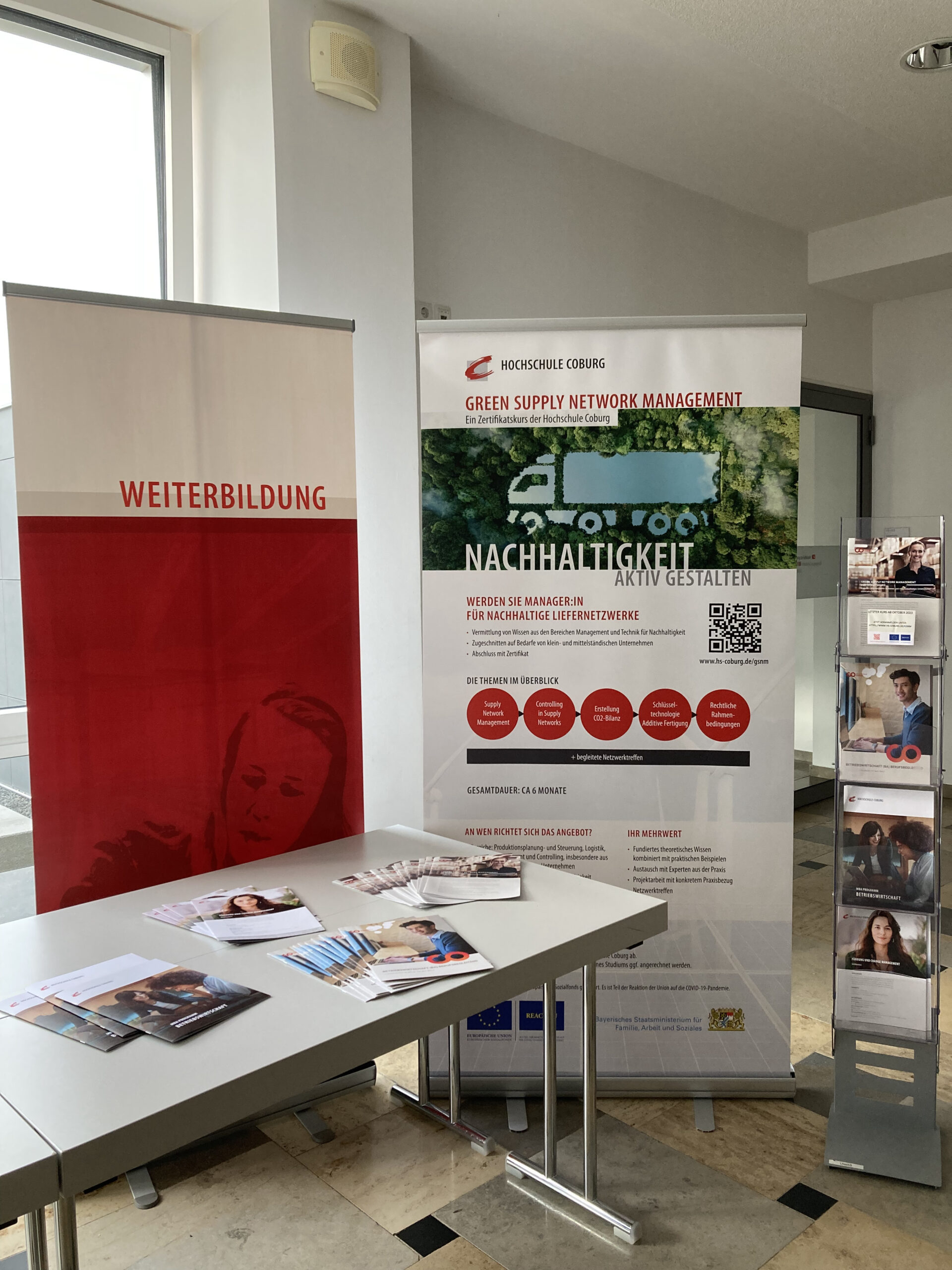 In einem Ausstellungsbereich werden auf einem Tisch vor zwei stehenden Bannern Broschüren präsentiert. Ein Banner ist rot und trägt den Text „Weiterbildung“, während das andere auf Deutsch für nachhaltiges Supply-Network-Management der Hochschule Coburg wirbt. Auf der rechten Seite befindet sich ein Prospektständer.