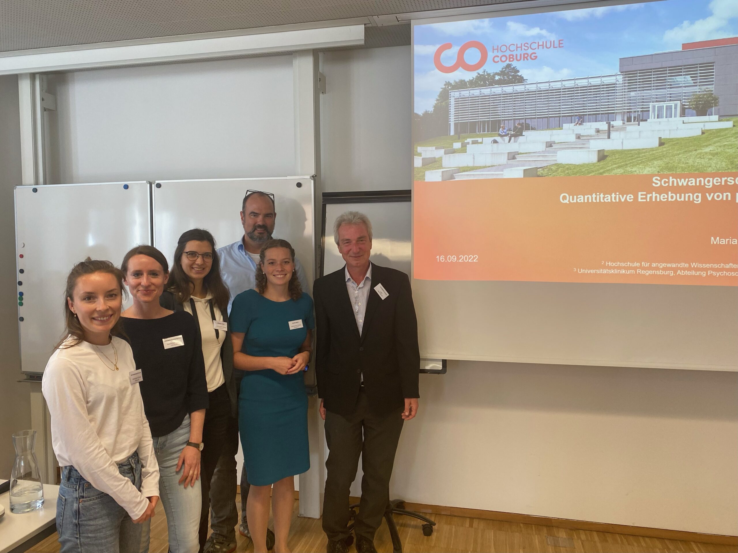 Eine Gruppe von sechs Personen steht in einem Hörsaal der Hochschule Coburg und posiert lächelnd vor einer Präsentationsfolie mit dem Titel „Schwangerschaft“. Im Hintergrund sind ein Whiteboard und eine Projektionsleinwand zu sehen.