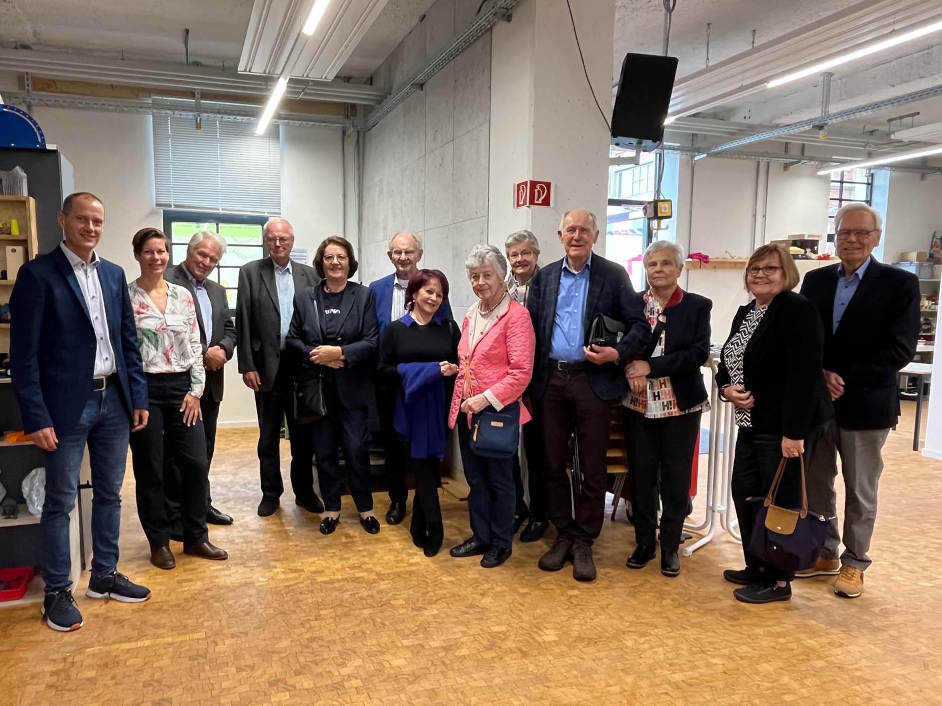 Eine Gruppe von zwölf Erwachsenen steht in einem Innenbereich der Hochschule Coburg auf einem Holzboden zusammen. Einige lächeln, sind leger gekleidet, im Hintergrund sorgen Regale und Decken im Industriestil für eine lockere und freundliche Atmosphäre.