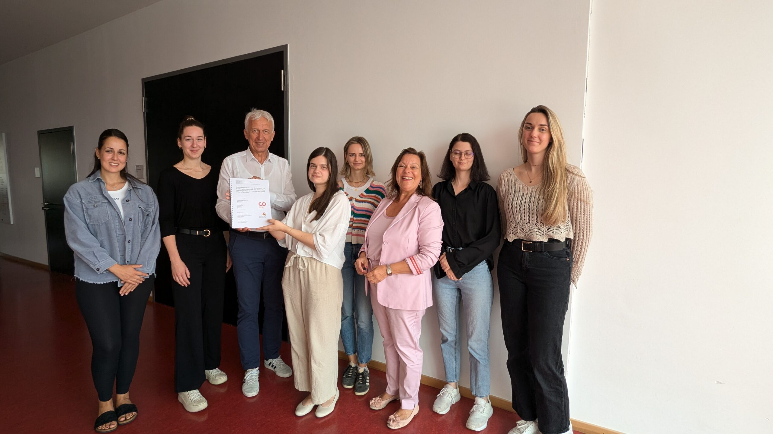 A group of nine people stands in a line against a white wall, perhaps at Hochschule Coburg. One person holds a document with a red logo. They are all smiling and dressed in casual and semi-formal attire, set indoors against the backdrop of a dark door.