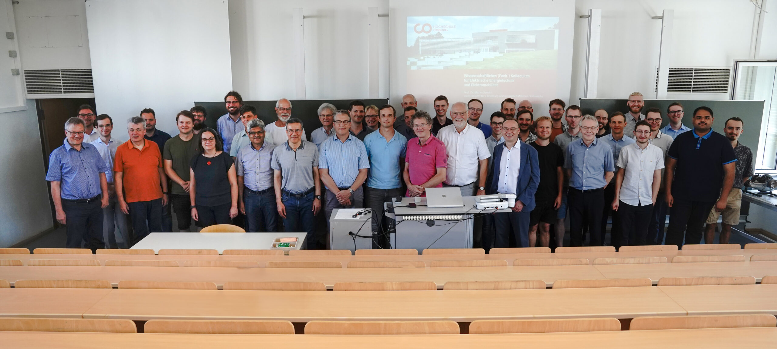 A group of people stands together in a lecture hall at Hochschule Coburg, posing for a photo. Some are smiling, while a presentation slide is displayed in the background. The hall features rows of wooden desks and crisp white walls.