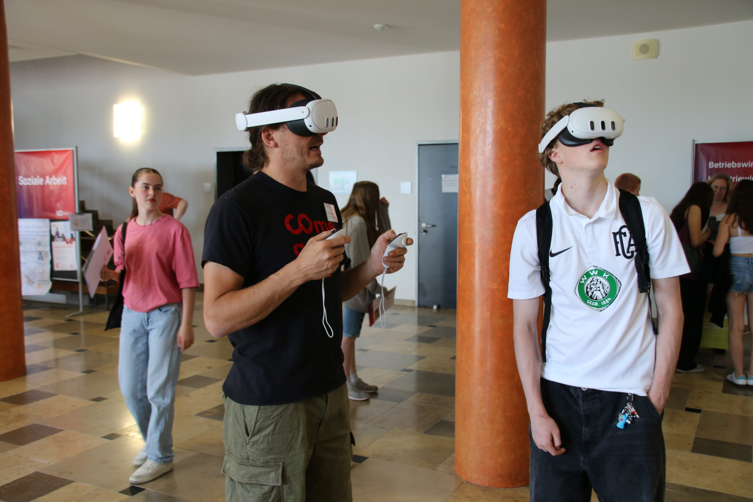 In a bright room at Hochschule Coburg, people wearing VR headsets interact with virtual content among columns. Observers on the tiled floor stand by, casually dressed and holding devices, immersed in this digital exploration.