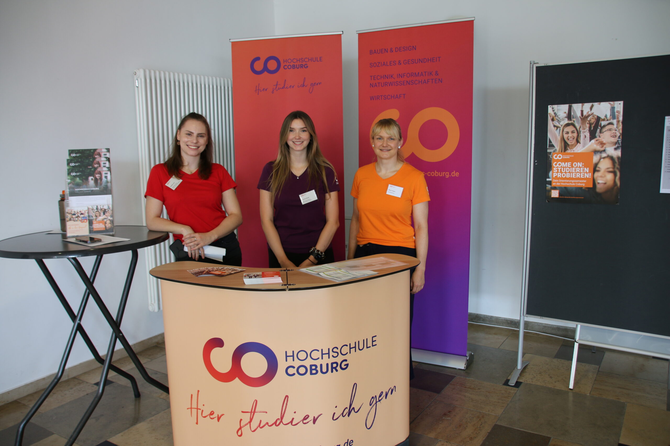 Behind the information booth for Hochschule Coburg, three people stand smiling, proudly wearing their name tags. The booth is adorned with brochures and a banner featuring the university's logo.