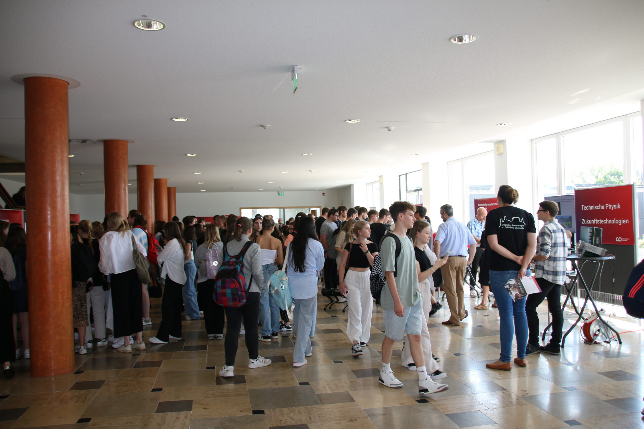 A large group of people gathered in a spacious, well-lit hall at Hochschule Coburg with wooden pillars and tiled floors. Some are conversing while others stand near a display with technology-related posters. Most are dressed casually.