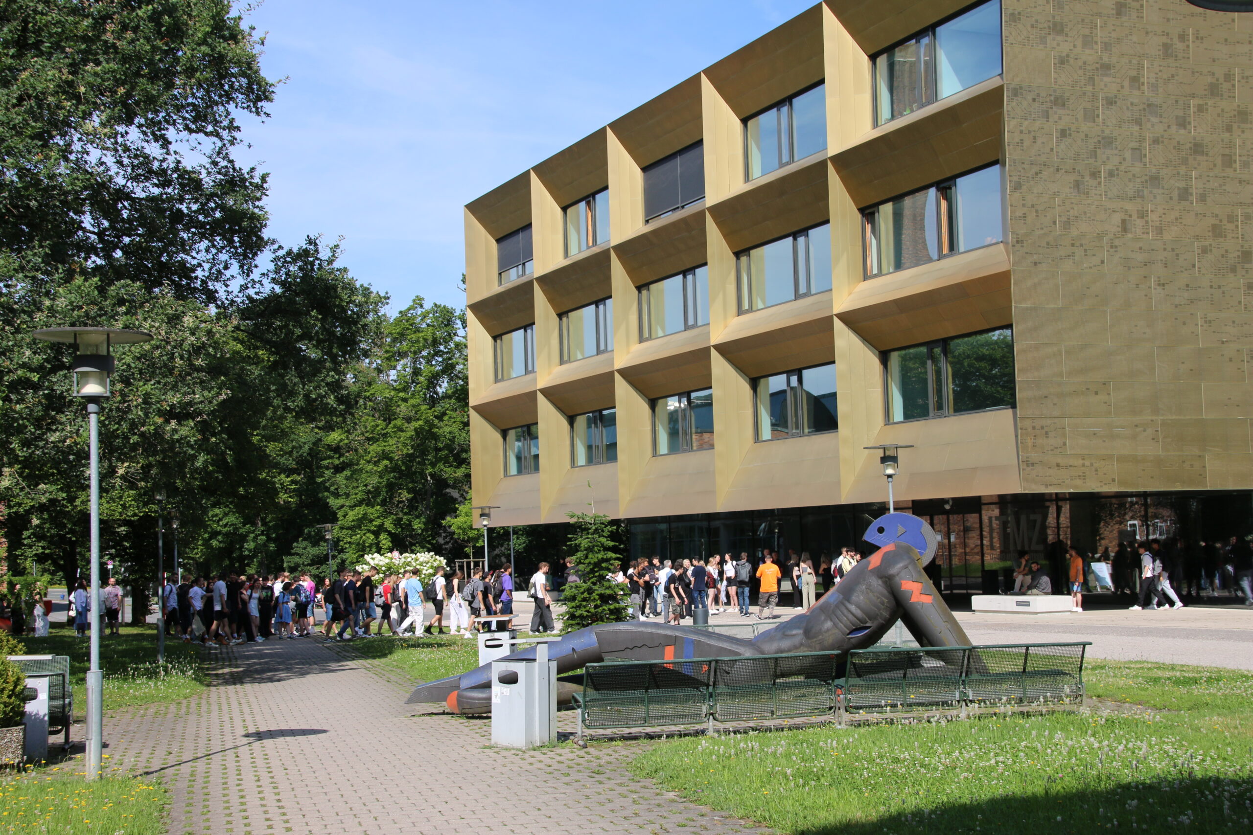 A large modern building with a gold-colored facade and geometric windows stands prominently, reminiscent of Hochschule Coburg’s architectural elegance. A group of people walk along the path, while in the foreground, a long, snake-like sculpture with colorful patterns weaves its charm. Trees grace the left side.