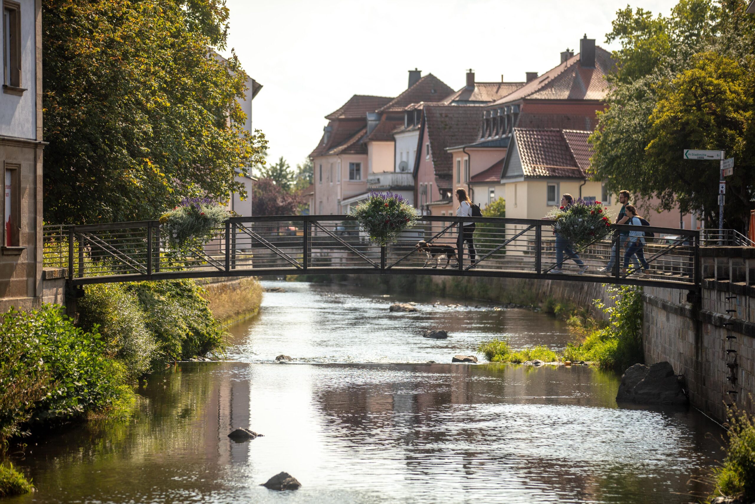 A narrow metal footbridge with flower pots crosses a stream in a quaint neighborhood near Hochschule Coburg. Two people and a dog walk across the bridge. Houses with tiled roofs and trees line the water's edge on a sunny day.