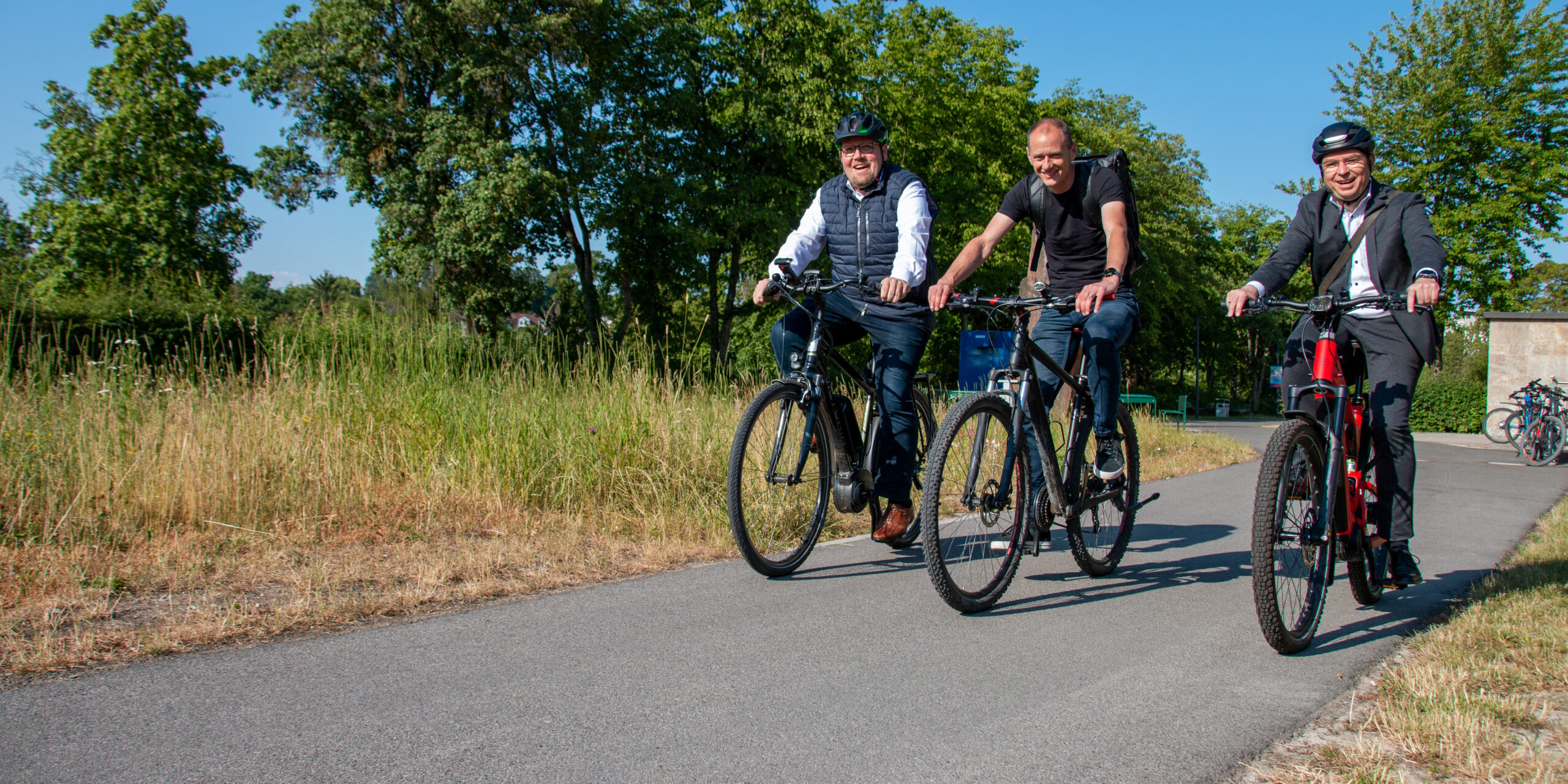 Drei Männer der Hochschule Coburg fahren mit dem Fahrrad auf einem gepflasterten Weg inmitten von Grün. Sie lächeln, tragen legere Kleidung und Helme und genießen den sonnigen Tag unter dem klaren blauen Himmel.