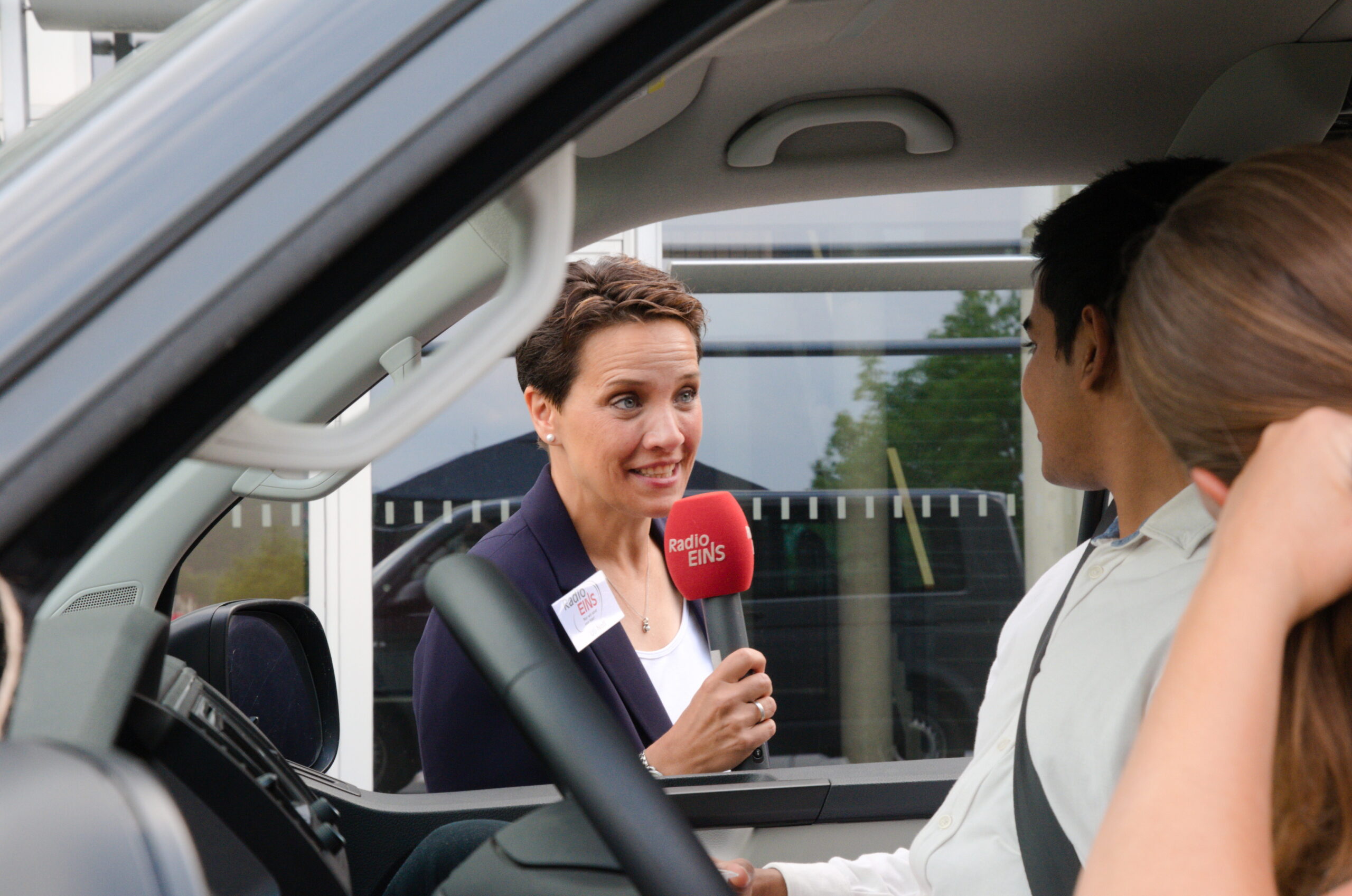 Eine Frau mit einem roten Mikrofon mit der Aufschrift „Radio EINS“ lehnt sich in ein Auto und spricht mit einem sitzenden Mann. Das Gespräch findet im Freien statt, in der Nähe des markanten Gebäudes mit den großen Fenstern der Hochschule Coburg.