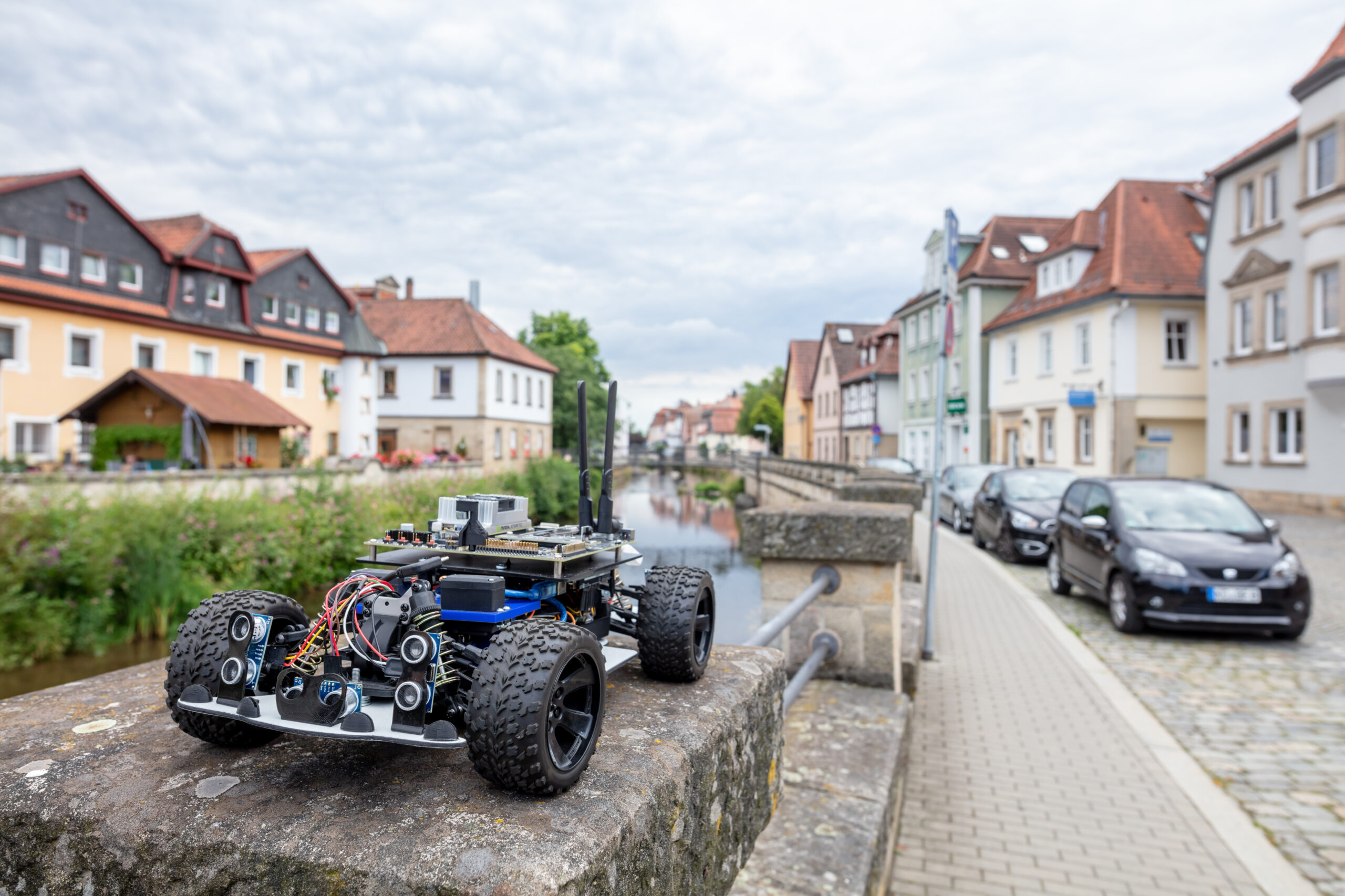 Ein kleines, robustes Roboterauto mit freiliegenden Kabeln und Elektronik steht auf einem steinernen Brückengeländer und bietet einen Blick auf die malerische Szenerie der charmanten Stadt der Hochschule Coburg, wo bunte Häuser und ein von parkenden Autos gesäumter Kanal die Kopfsteinpflasterwege unter einem bewölkten Himmel zieren.